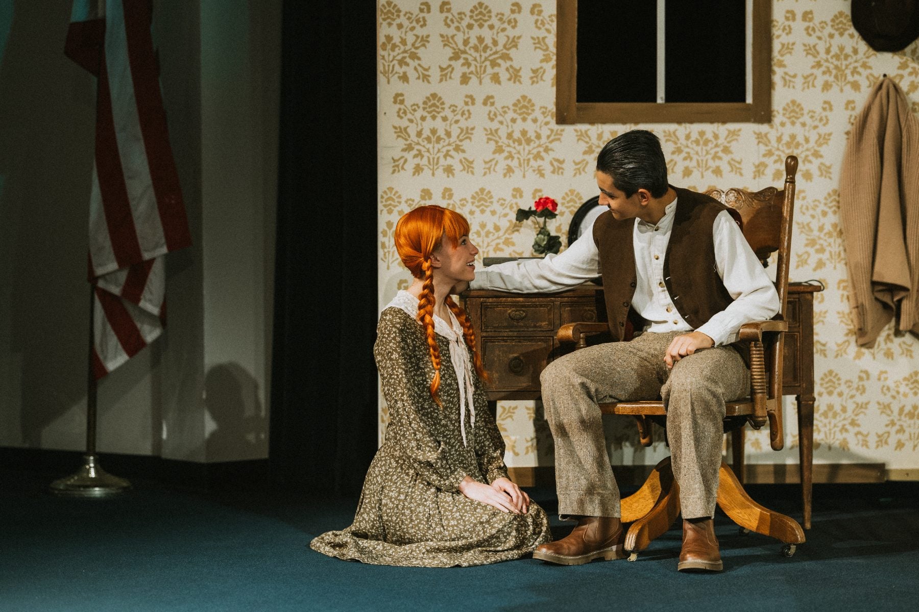 A student kneels beside a seated classmate near an American flag on set