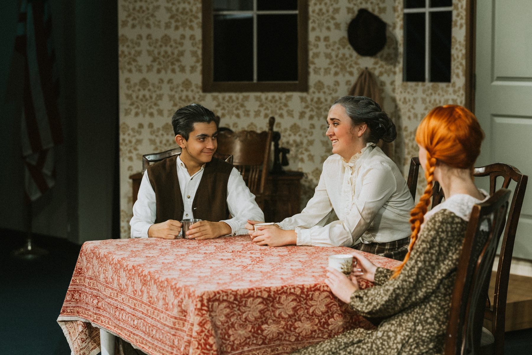 Three students share a conversation around a table in a staged parlor scene