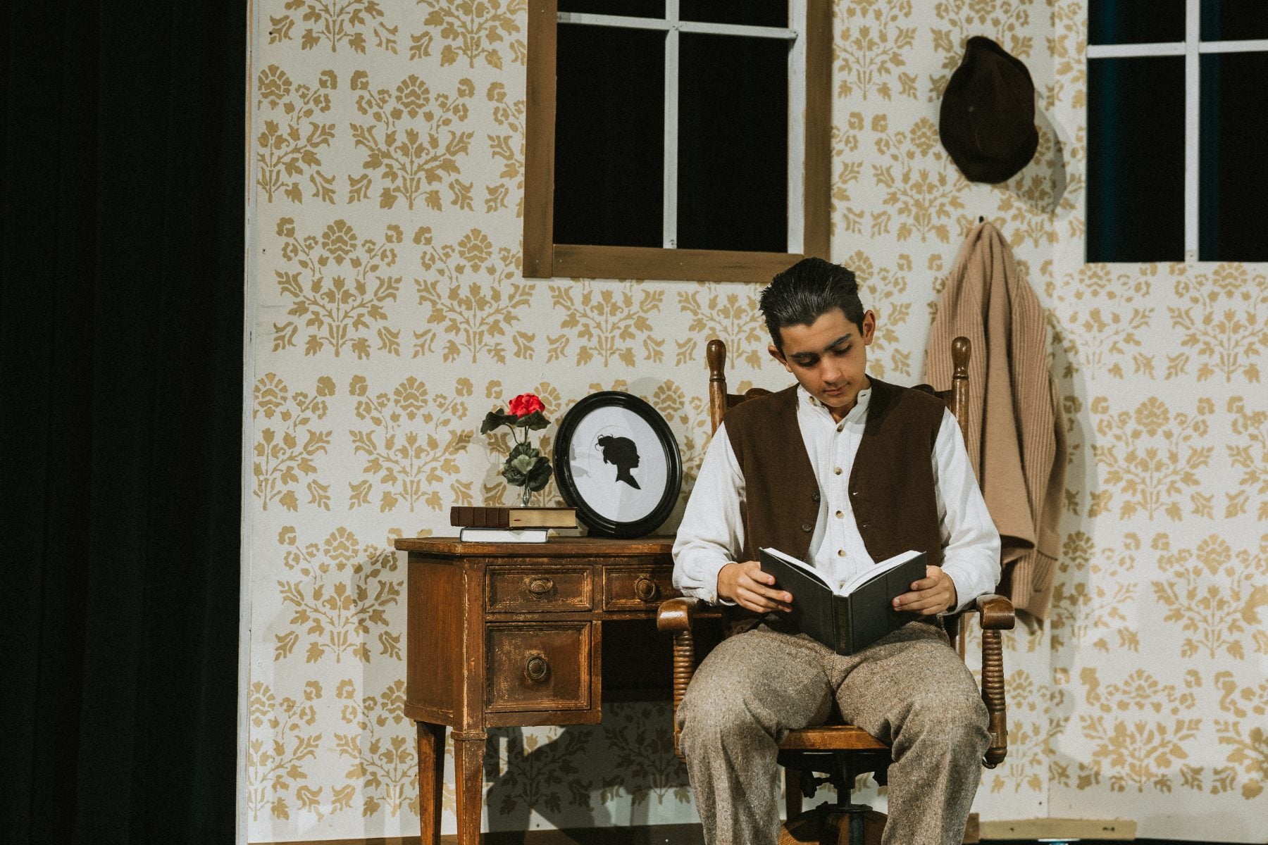 A student in costume reads a book while seated in a rocking chair on set