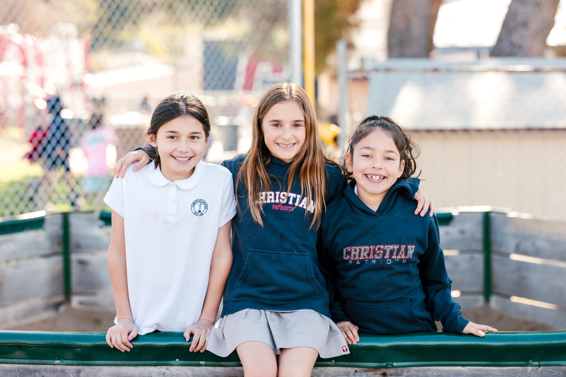 Three girls sit on a bench with arms around each other, smiling