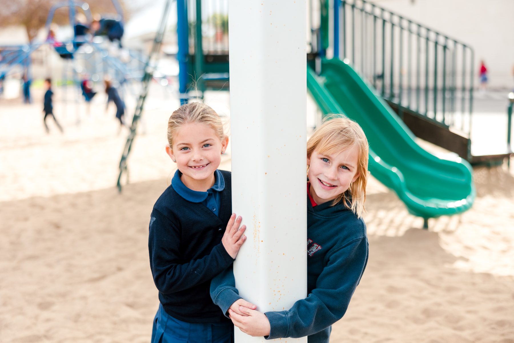 Two girls peek around a playground pole near the slide, smiling