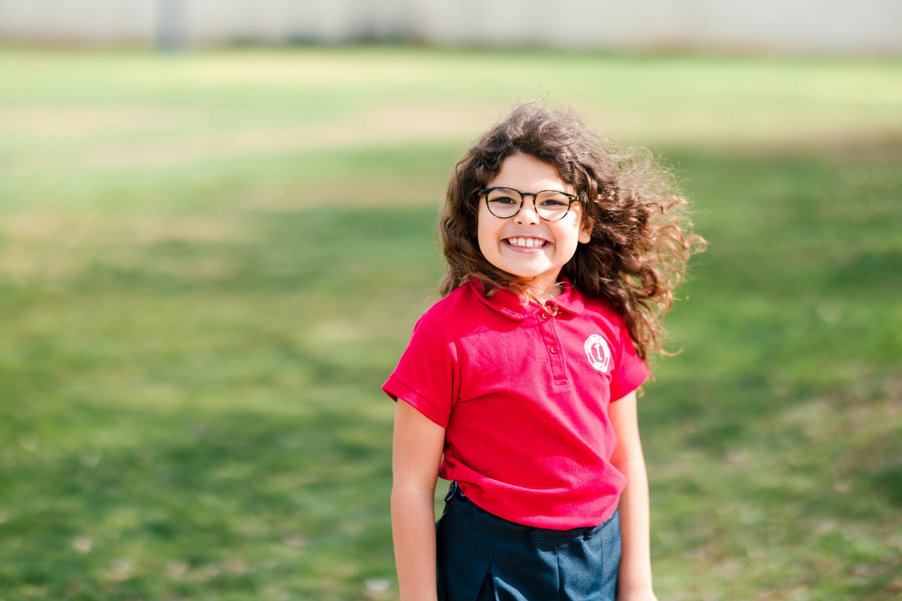 A smiling kindergarten girl in a red polo stands outside on the grass