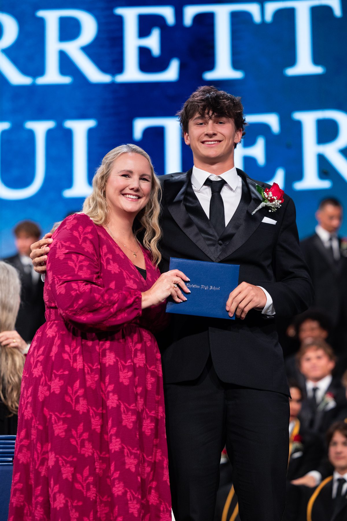 A woman in red embraces a graduate holding his diploma on stage