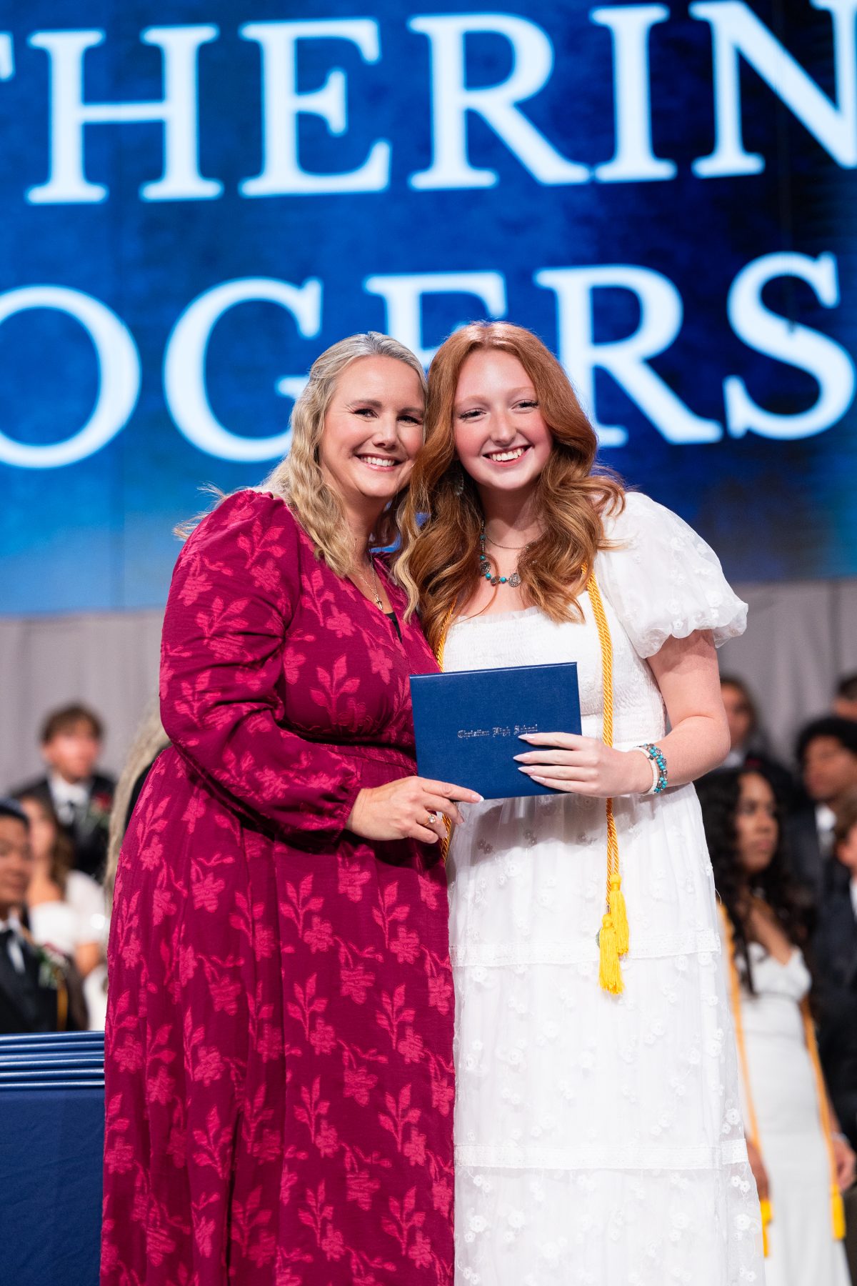 A teacher presents a diploma to a graduate beside a screen reading her name