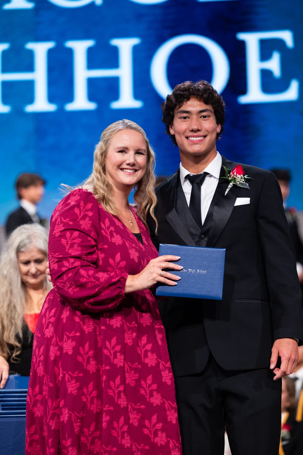 A staff member gives a diploma to a male graduate wearing a red rose
