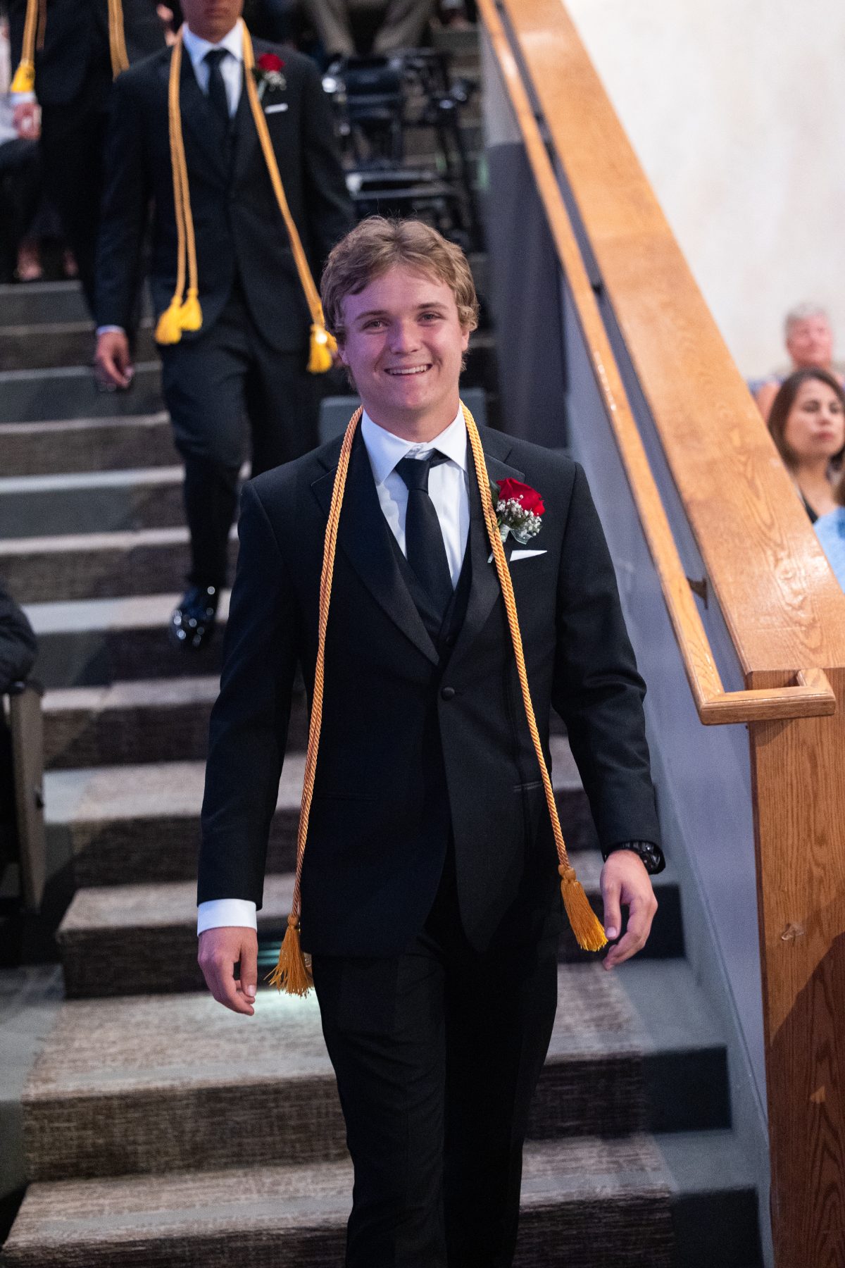 A graduate in a tuxedo with gold cords walks down the stairs at commencement