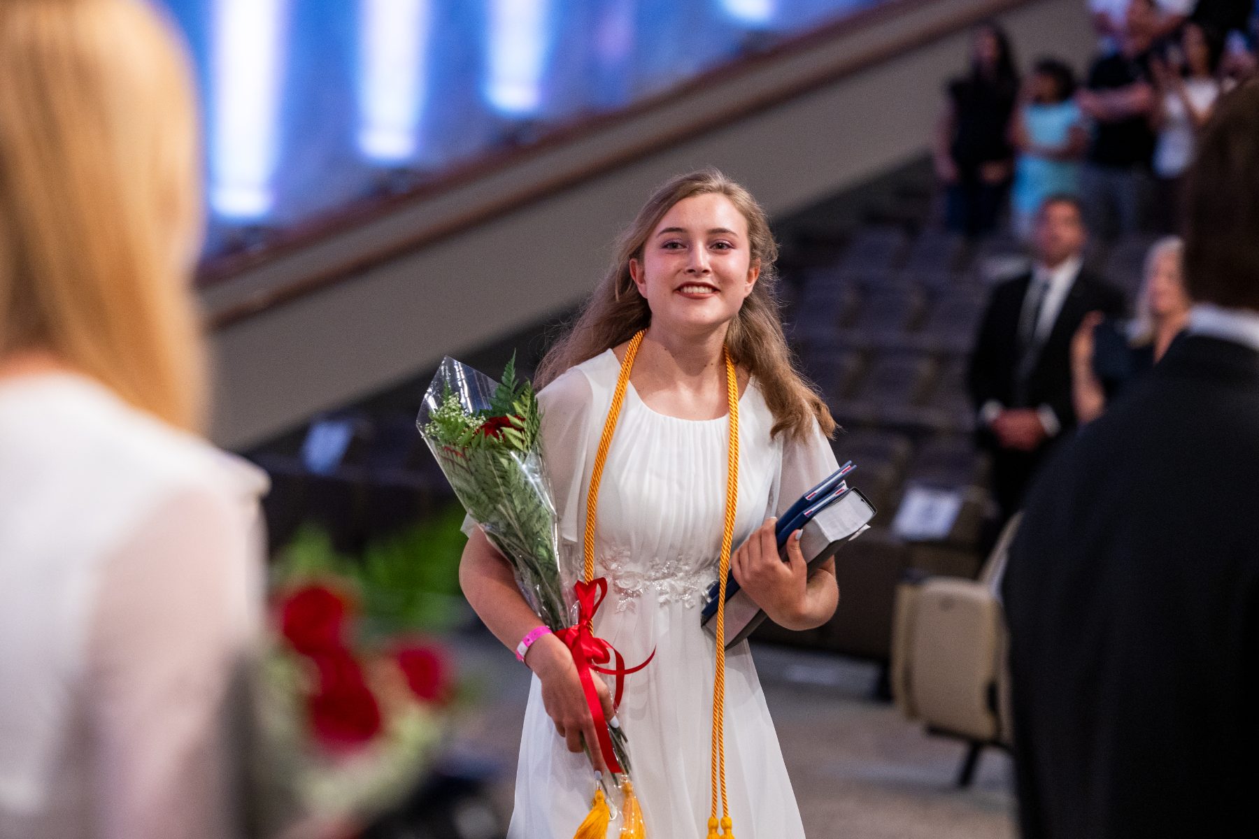 A graduate in a white dress carries flowers and a diploma across the stage