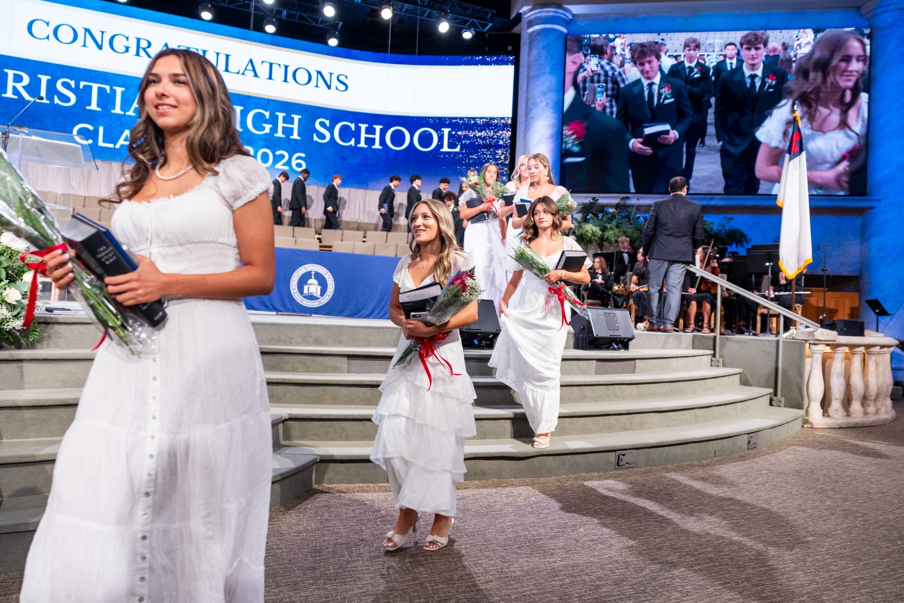 Graduates in white dresses descend stage steps under a Class of 2026 congratulations banner