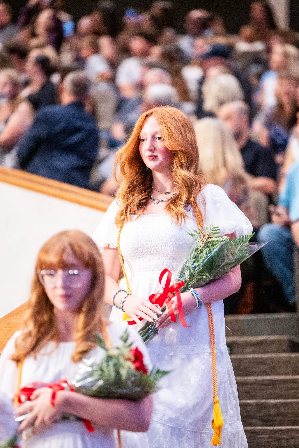 Two graduates in white dresses descend stairs holding bouquets