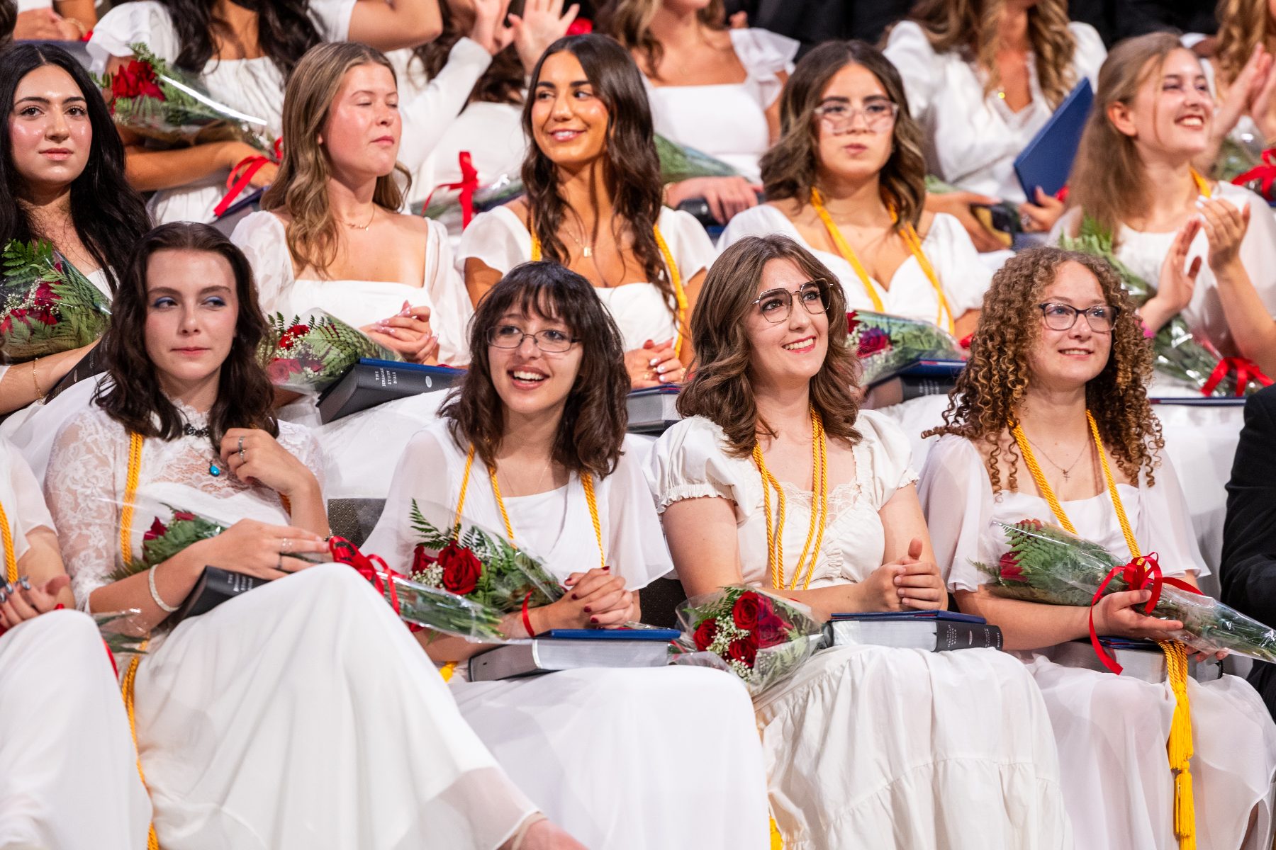 Graduates in white dresses hold roses while seated in the audience at commencement