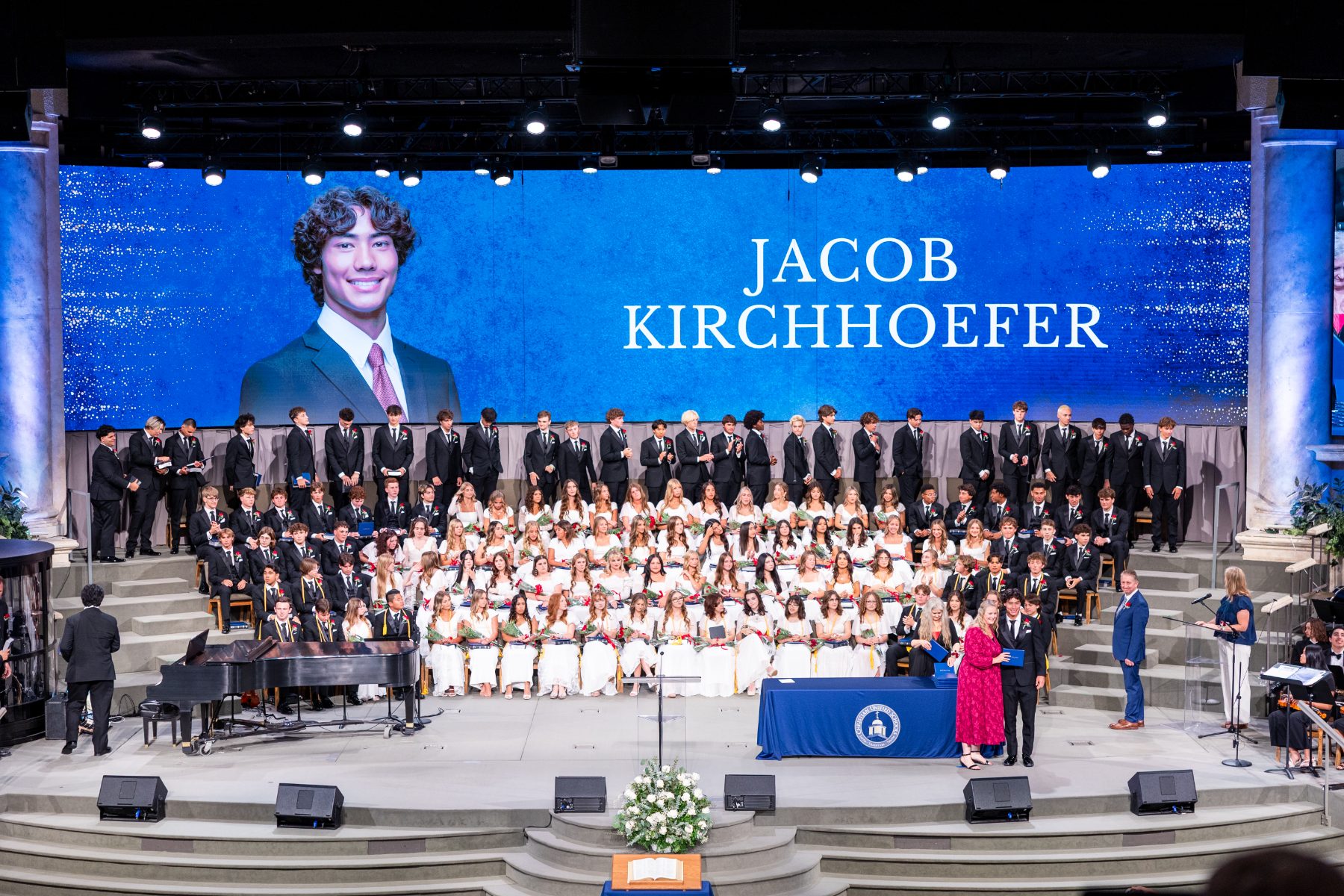 Graduates and choir stand on stage as a student's photo and name are displayed