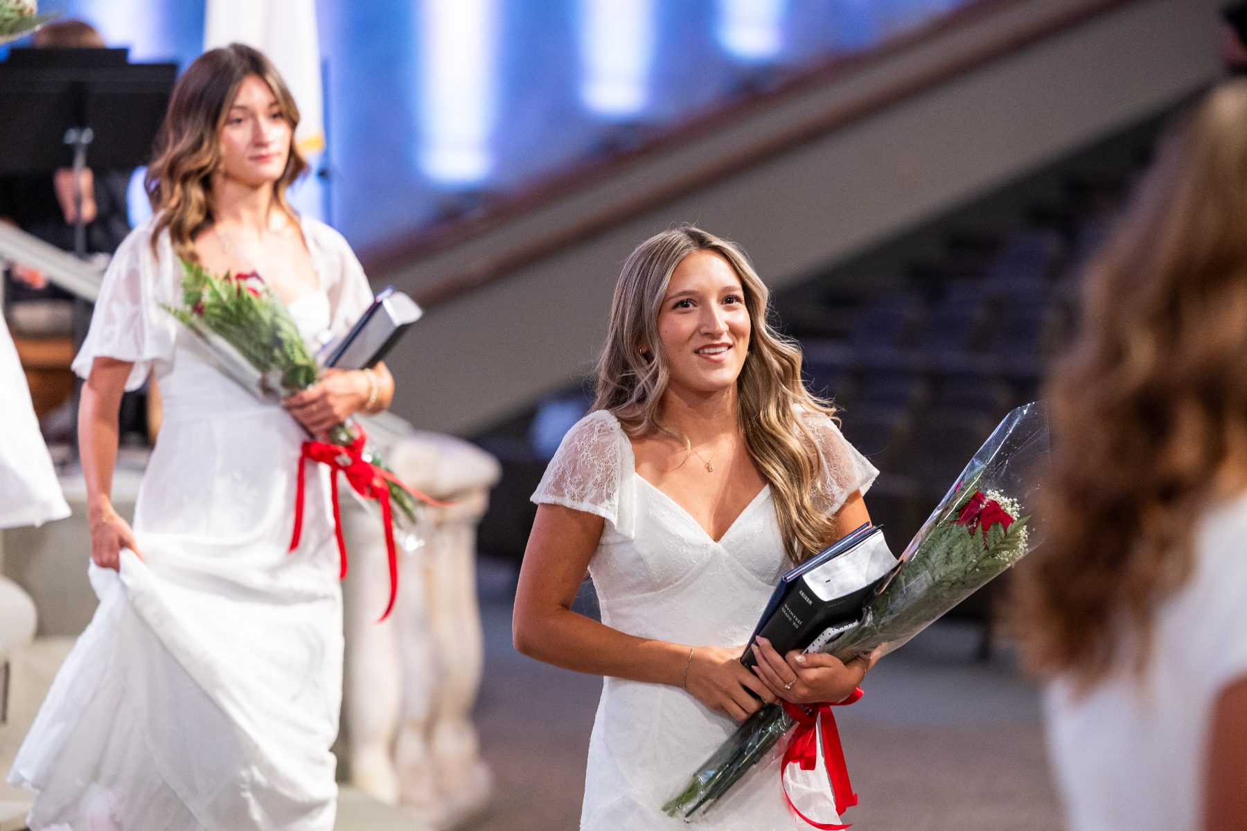 Two graduates in white dresses walk together holding flowers and diplomas