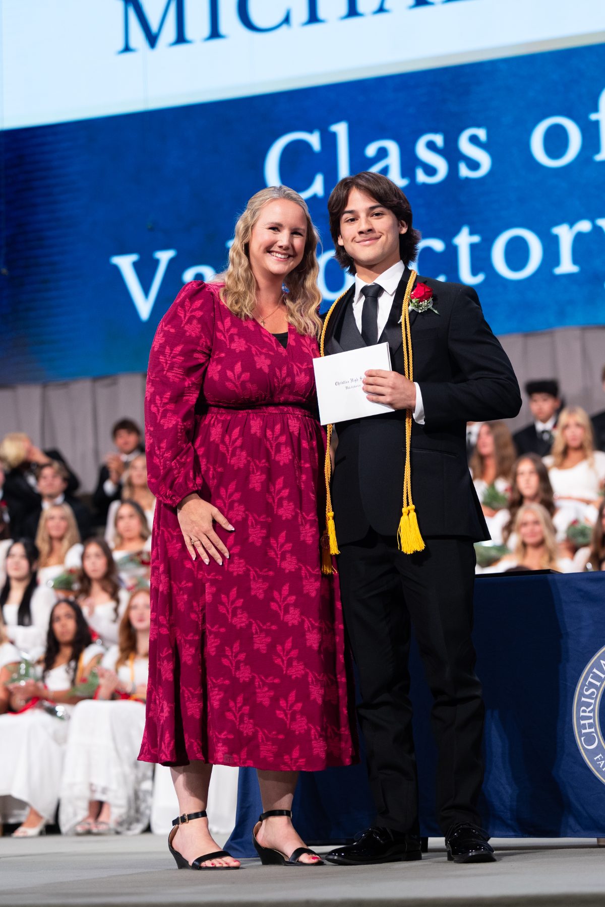 A student in cap-free attire receives a certificate from a woman on stage for valedictory honors