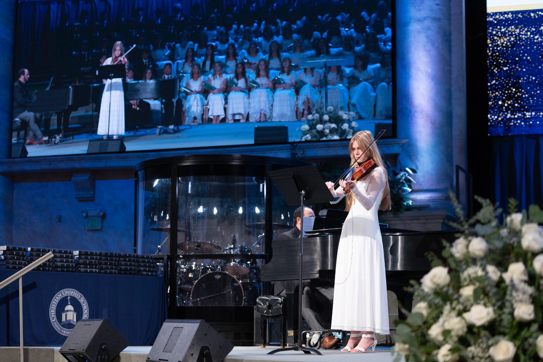 A student performs a violin solo on stage as a choir sings behind her