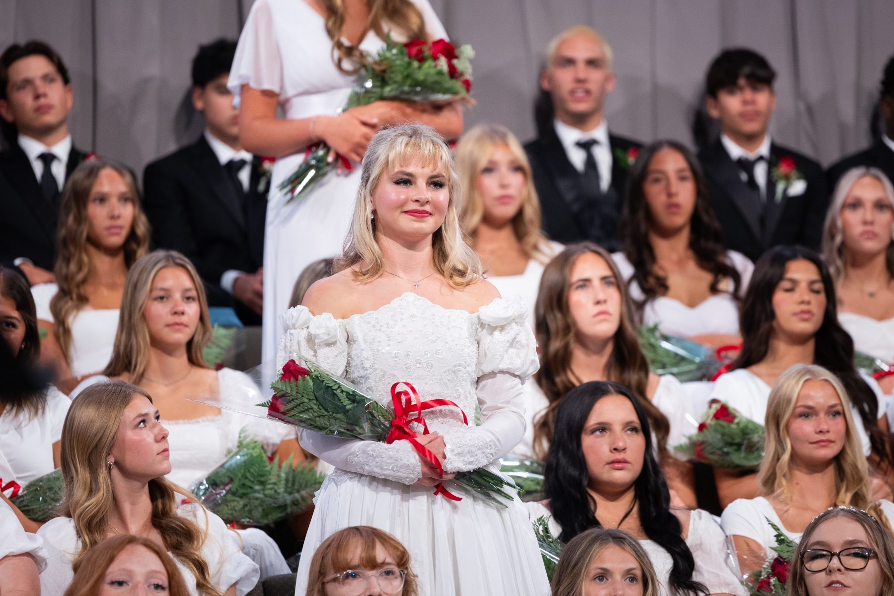 A graduate in white gown holds red roses among seated Christian High School classmates.