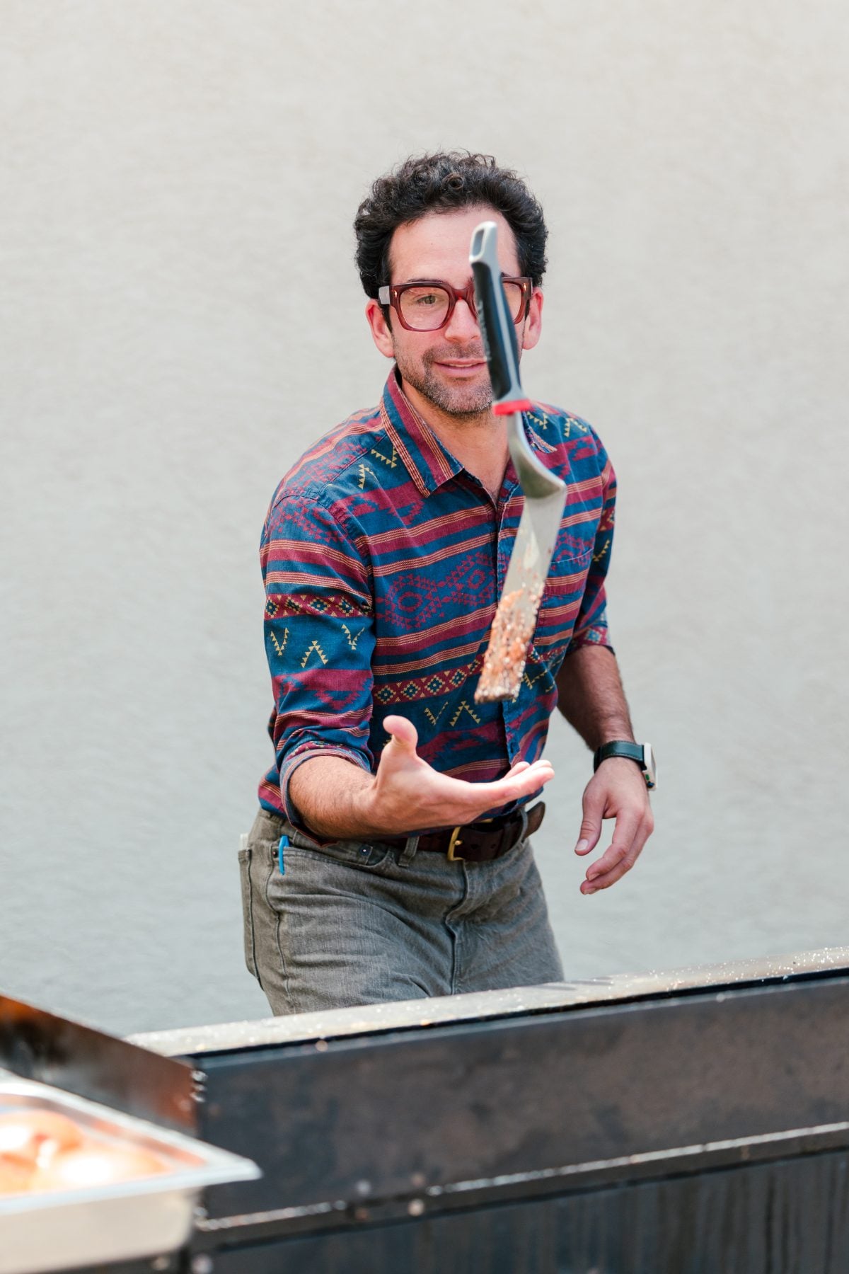 The BBQ cook flipping a grilling tool in the air outdoors