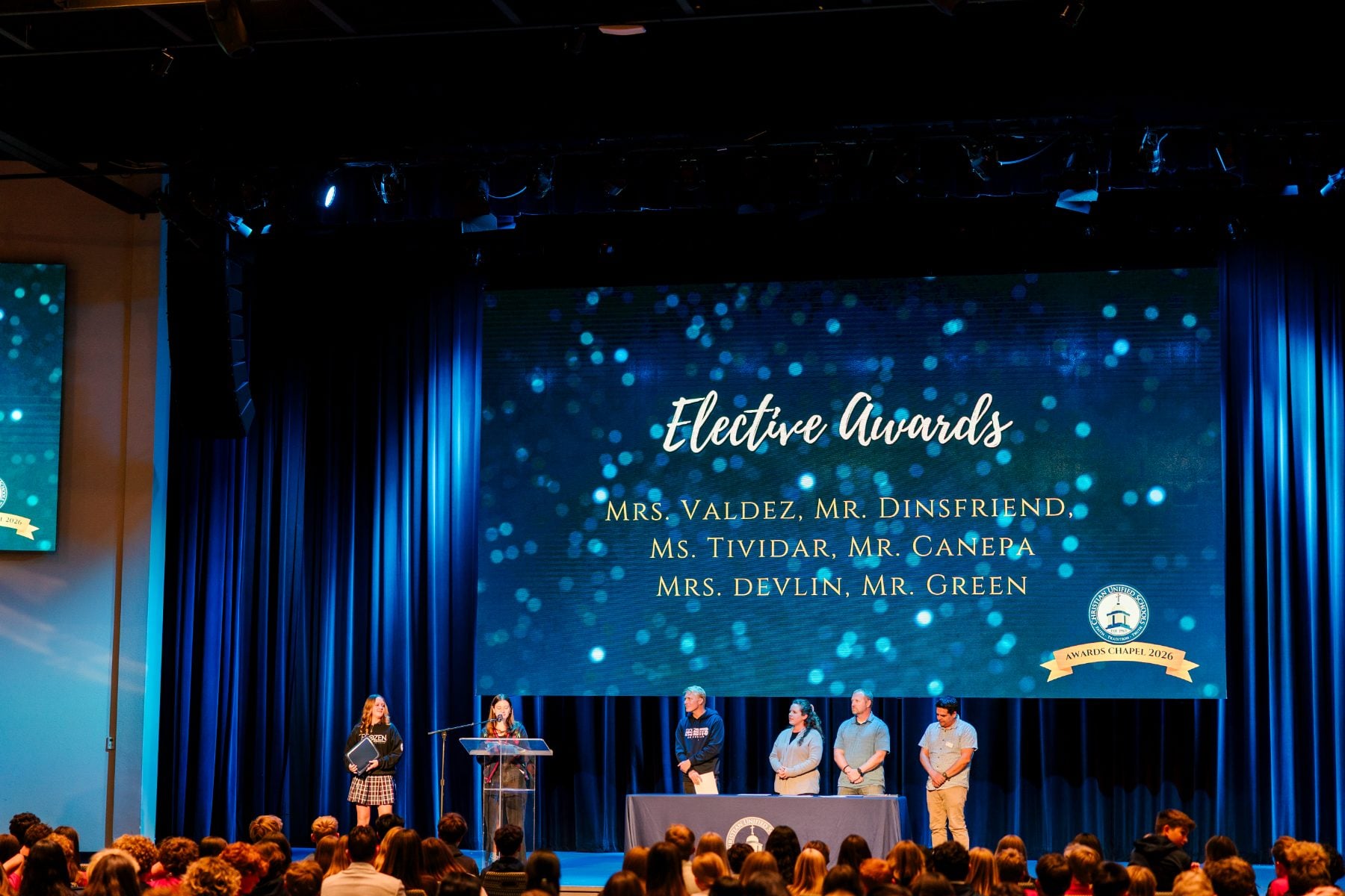 Staff stand on stage as the screen displays Elective Awards category winners.