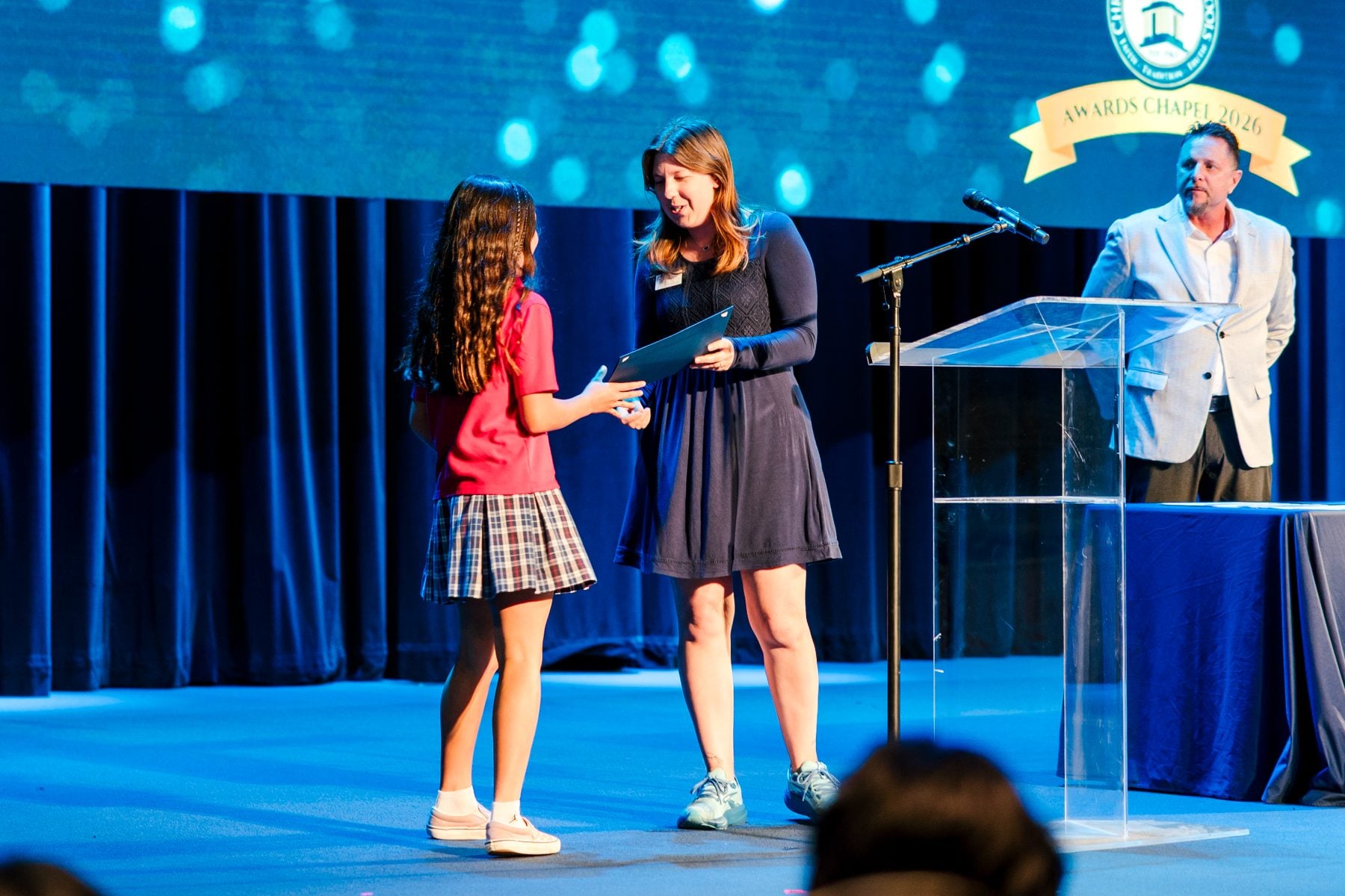 A young student in a plaid uniform accepts an award from her teacher.