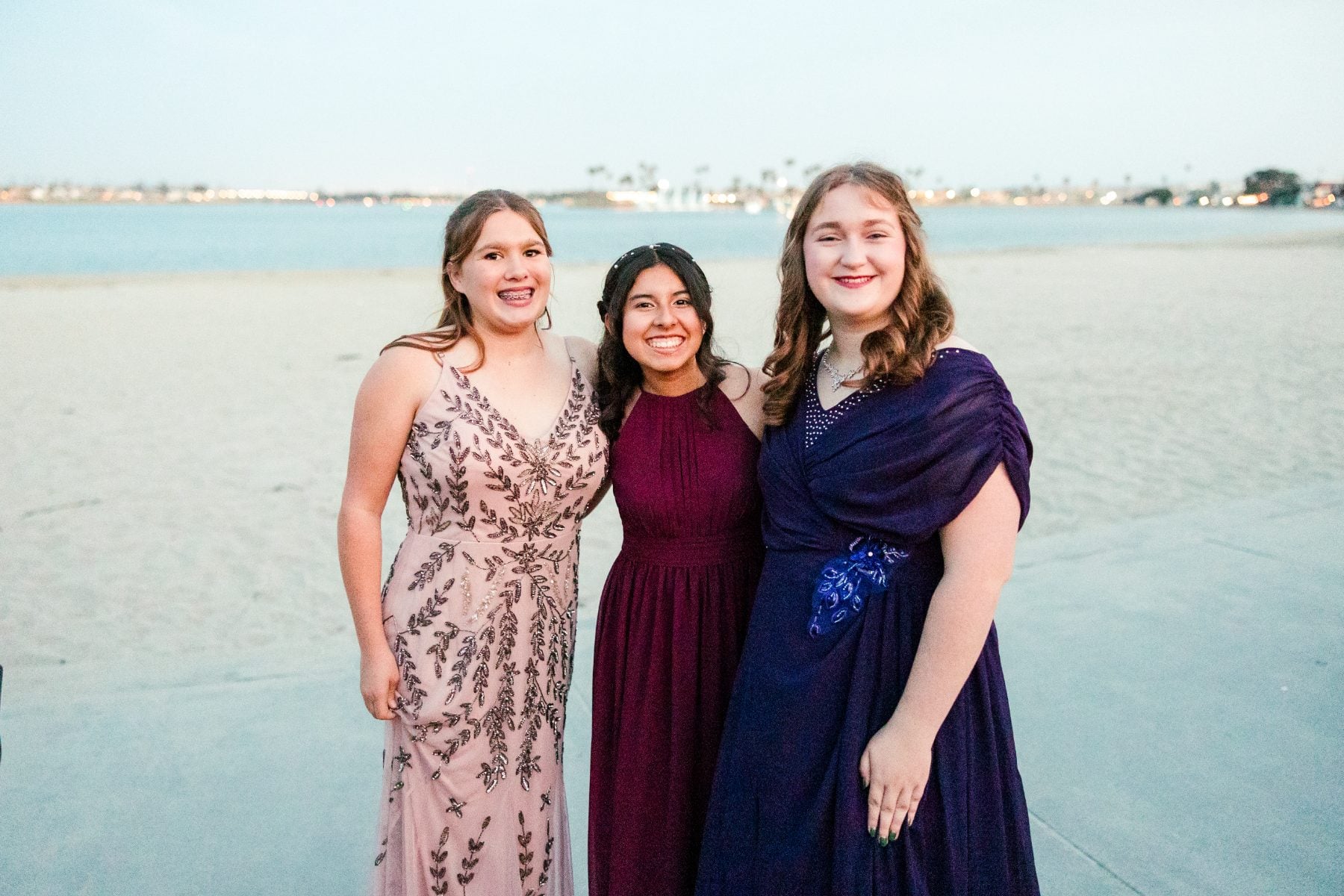 Three students in formal dresses smile together on the beach