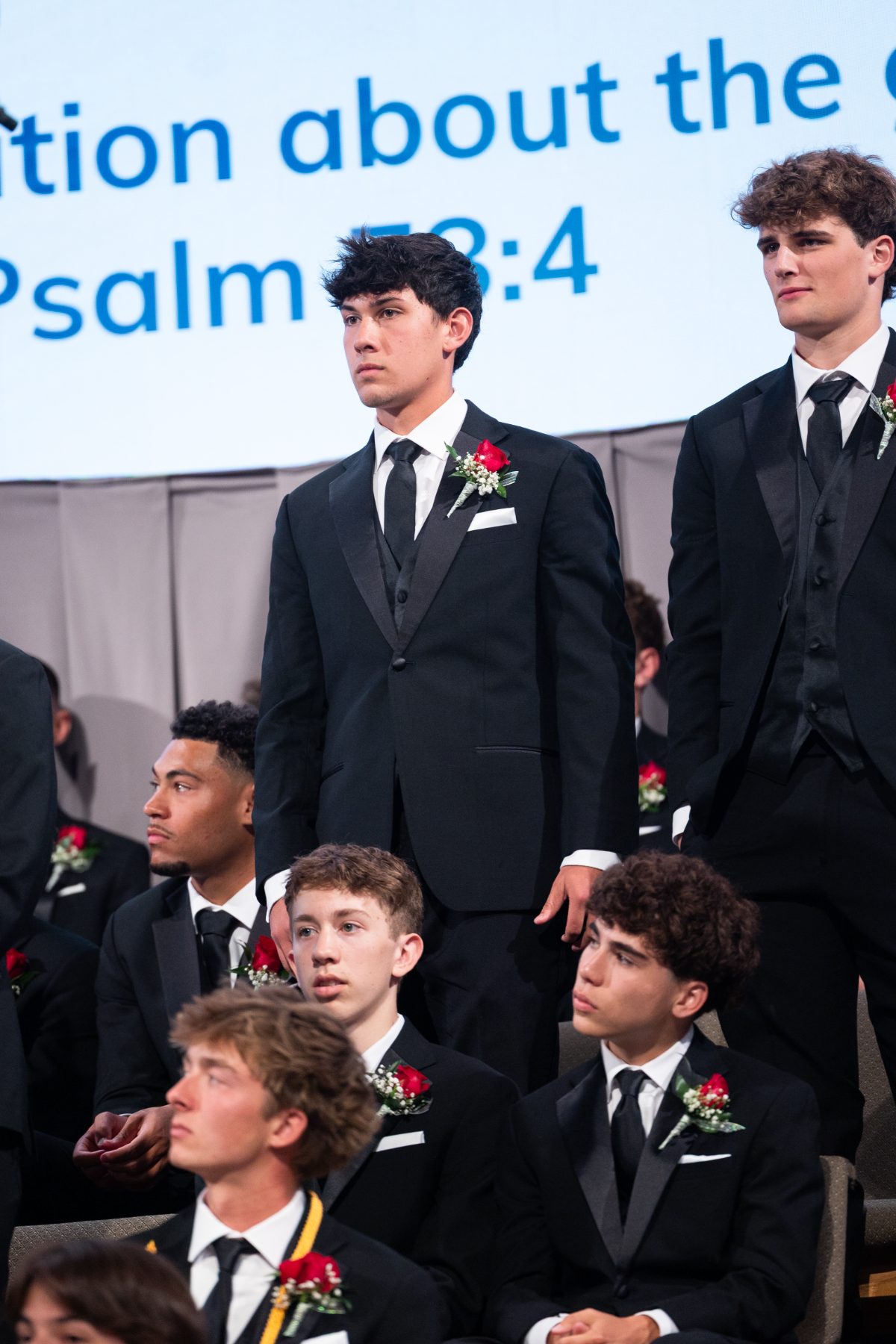 Students stand solemnly on stage as a Psalm verse displays behind them at graduation
