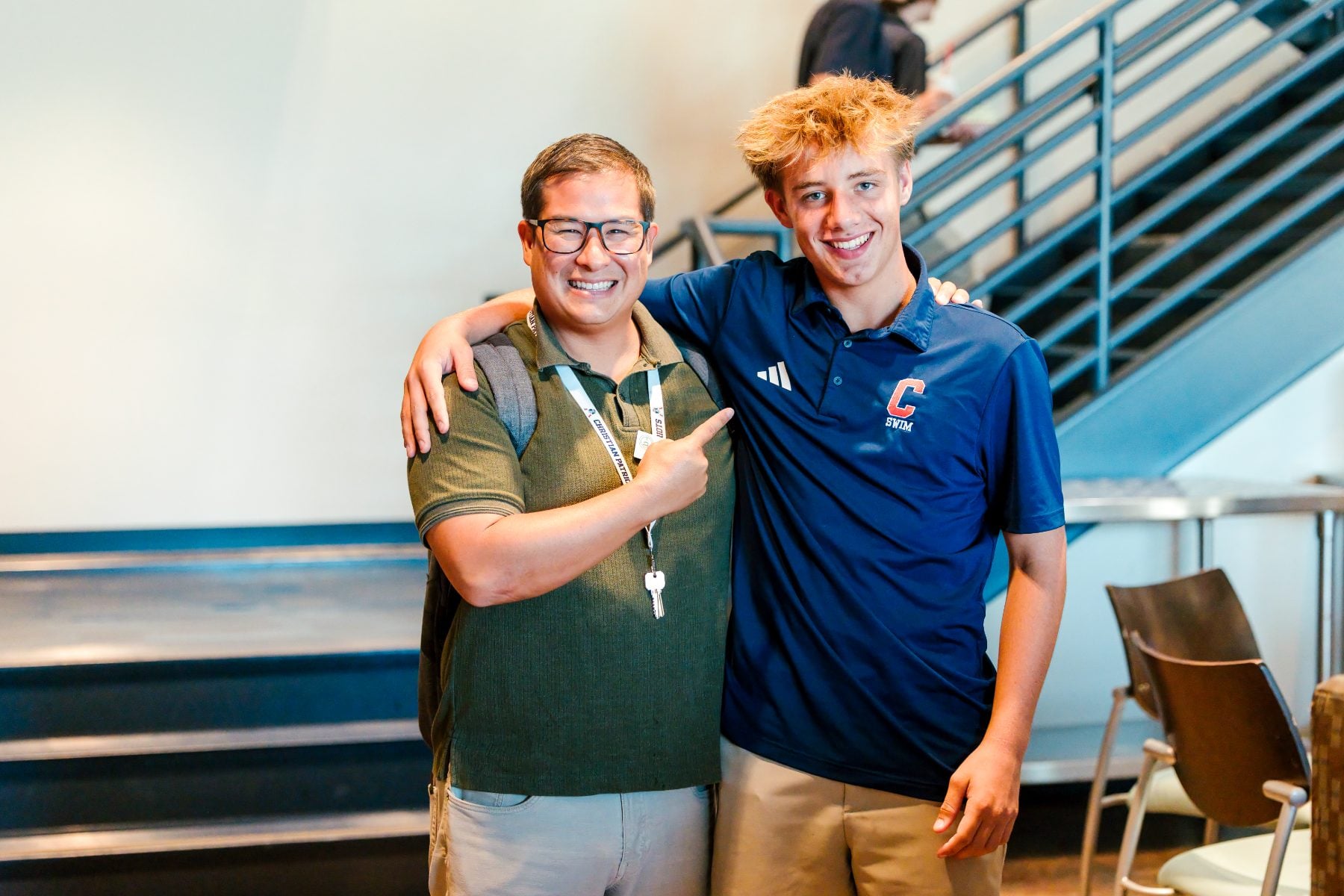 A teacher and student pose together near a staircase indoors