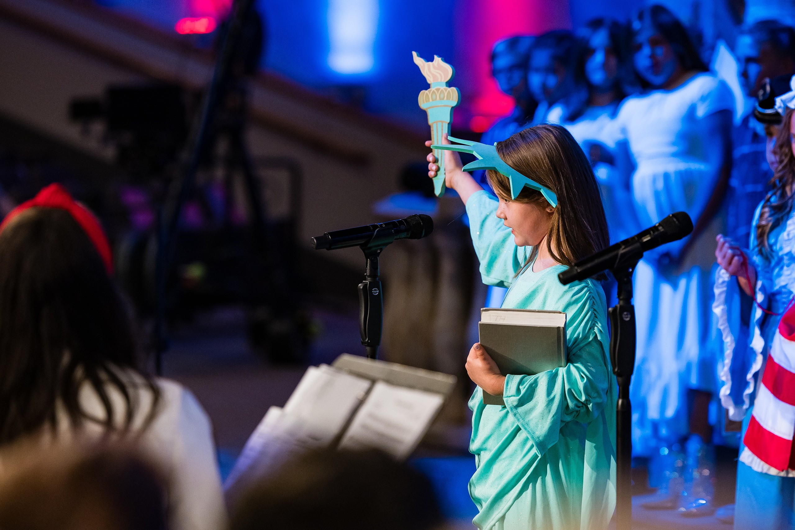 Christian Elementary East Celebrates Spring with “One Nation Under God” — 06 Student in green Statue of Liberty costume holding torch prop at microphone