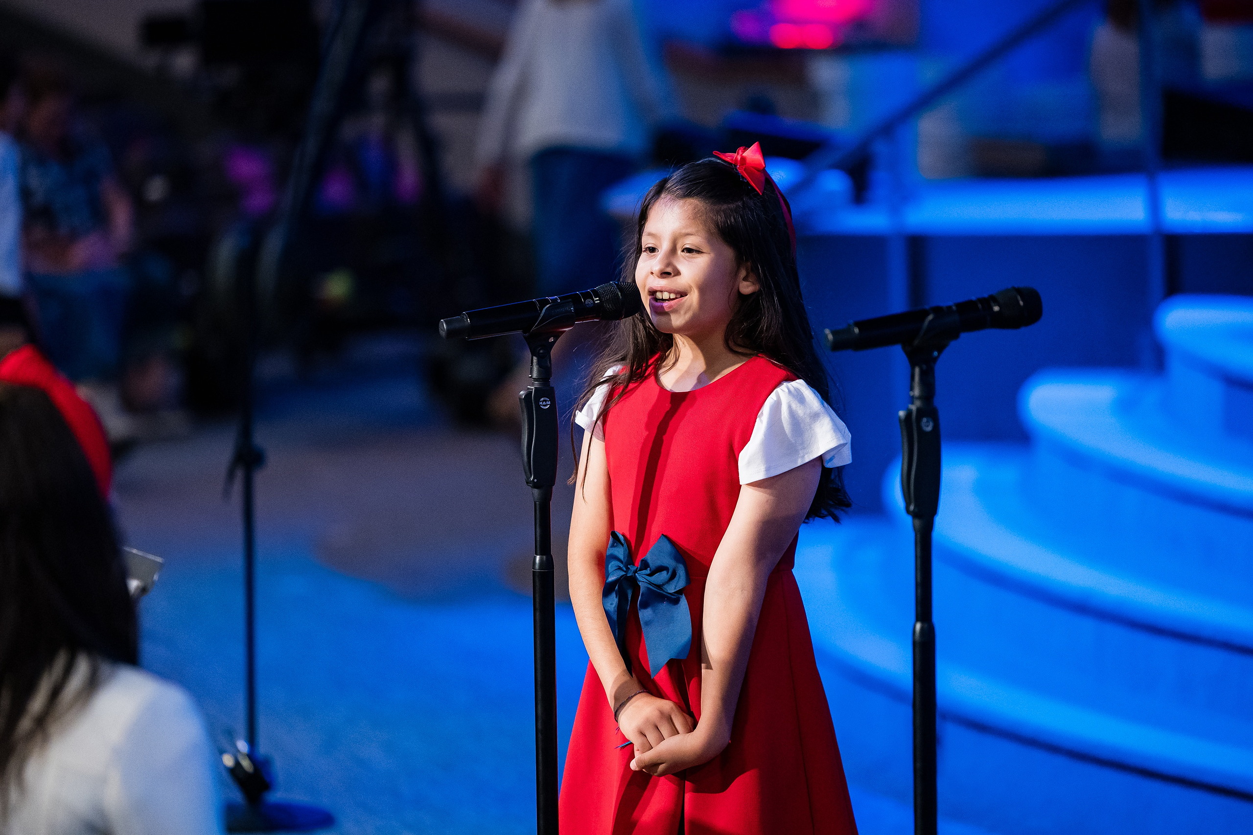 Christian Elementary East Celebrates Spring with “One Nation Under God” — 16 Student in red dress with white sleeves singing solo at microphone