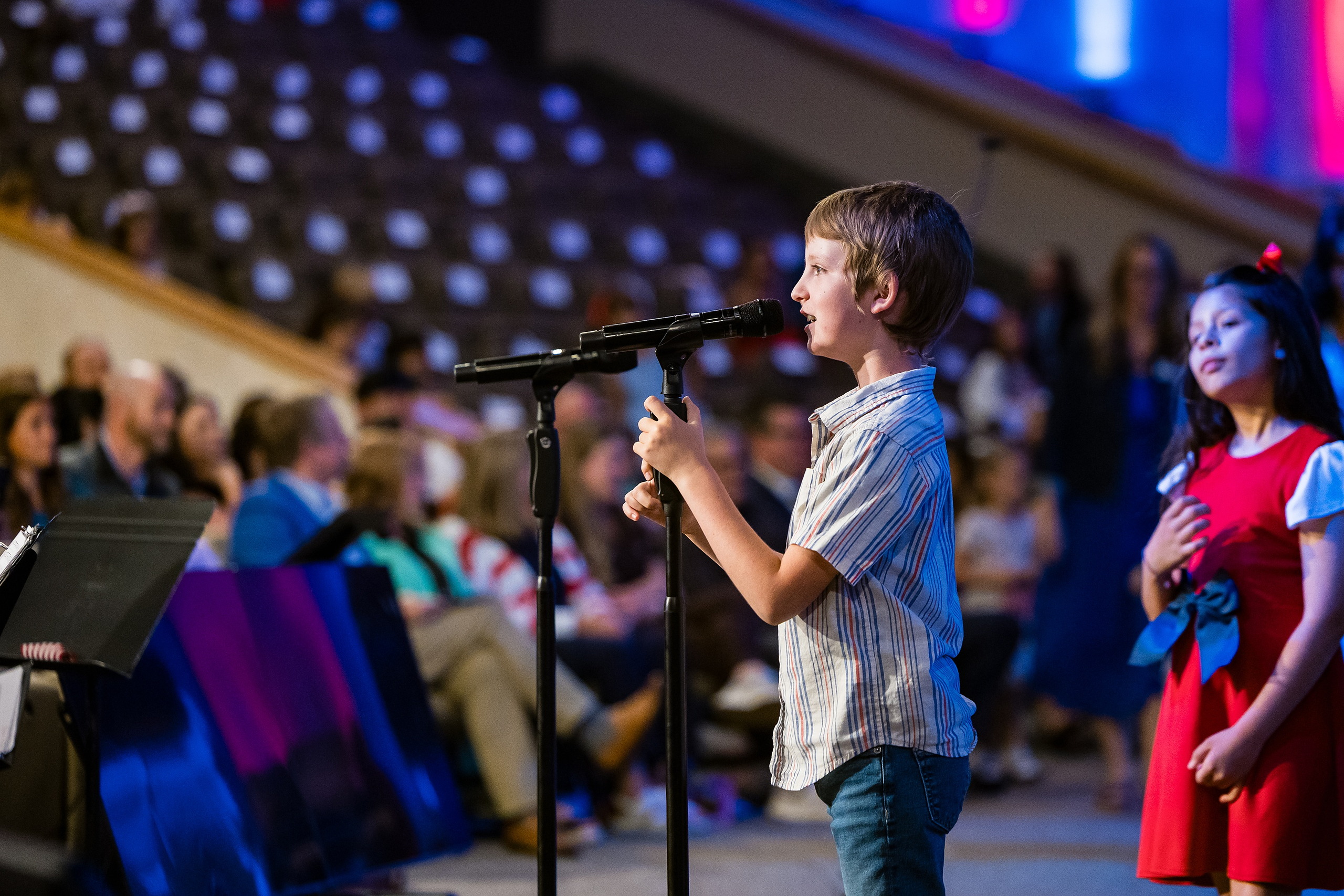 Christian Elementary East Celebrates Spring with “One Nation Under God” — 18 Young student in light striped shirt singing at microphone to audience