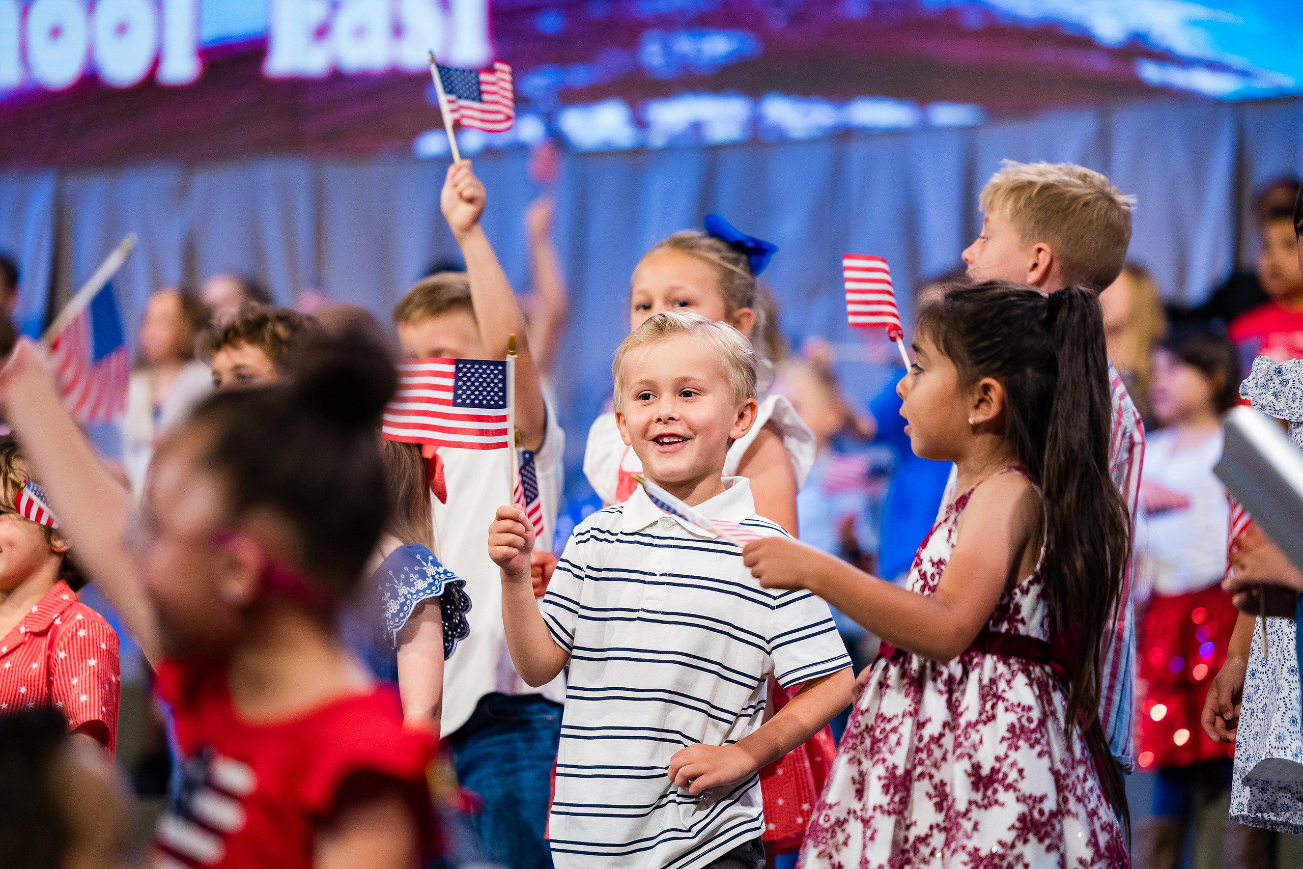 Christian Elementary East Celebrates Spring with “One Nation Under God” — 21 Children of various ages with small American flags smiling on stage