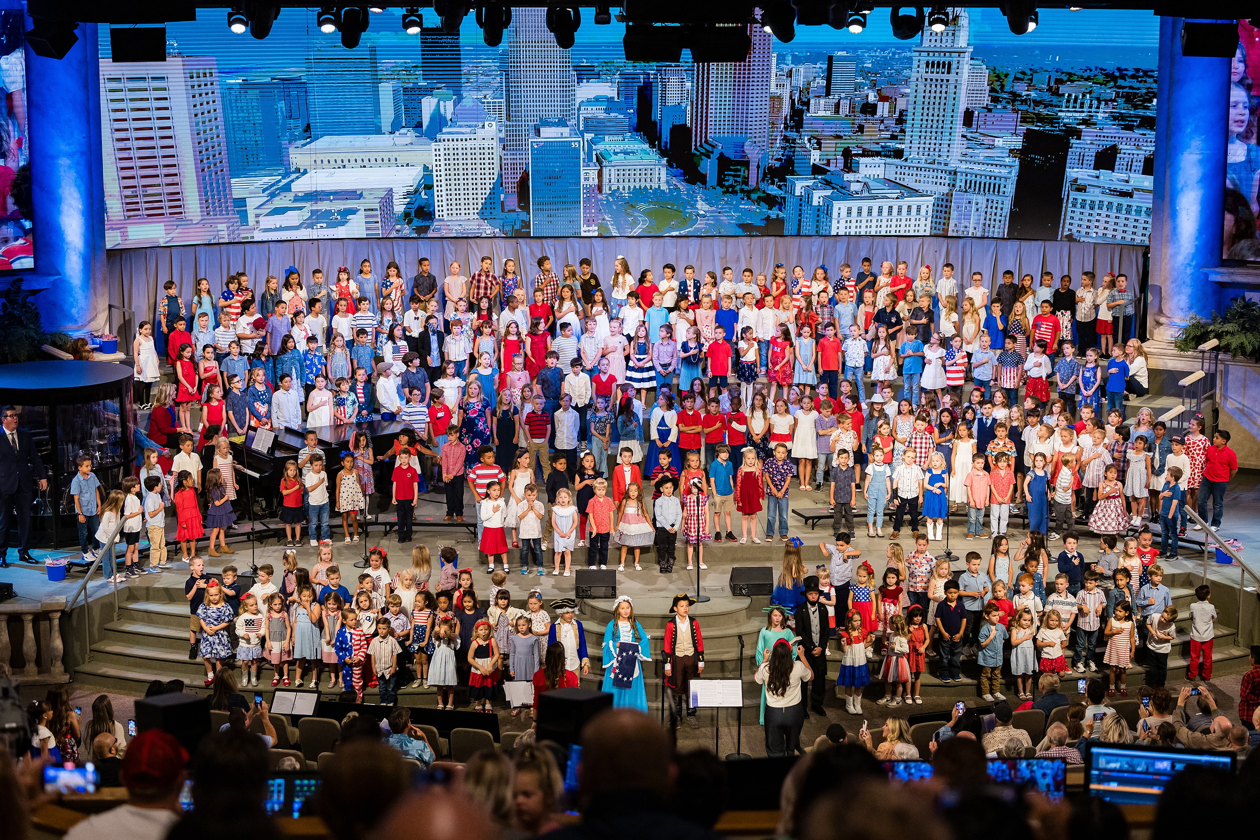 Christian Elementary East Celebrates Spring with “One Nation Under God” — 25 Full cast of all students arranged on stage with cityscape backdrop