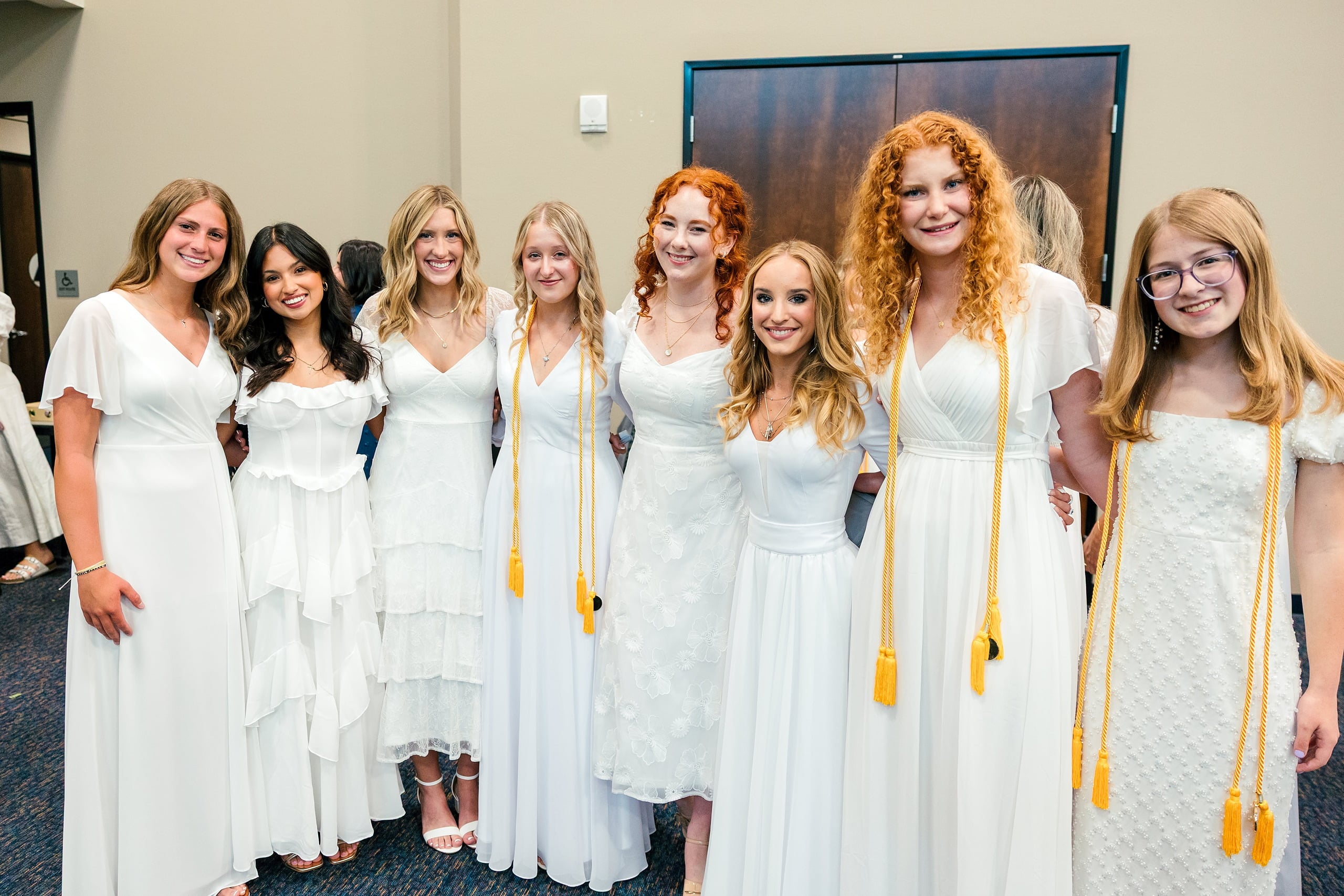 Christian High School Commencement 2025: Rise Up — 02 Eight female graduates in white dresses with gold cords stand in a row indoors