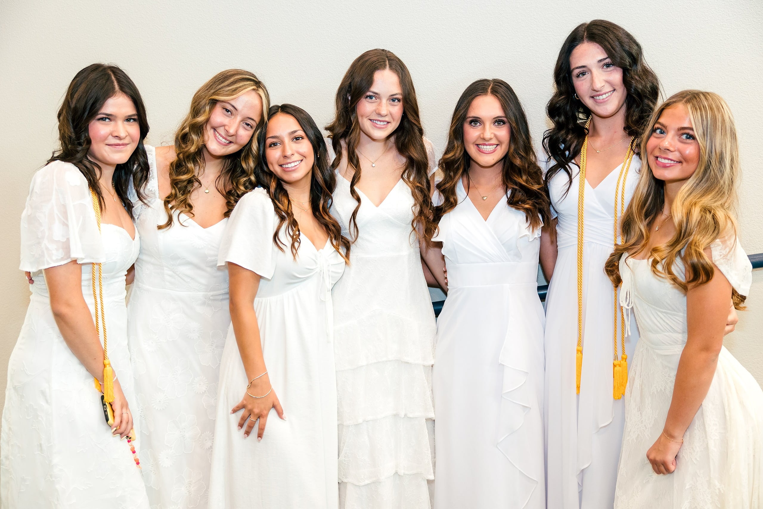 Christian High School Commencement 2025: Rise Up — 05 Seven female graduates in white dresses stand together indoors celebrating