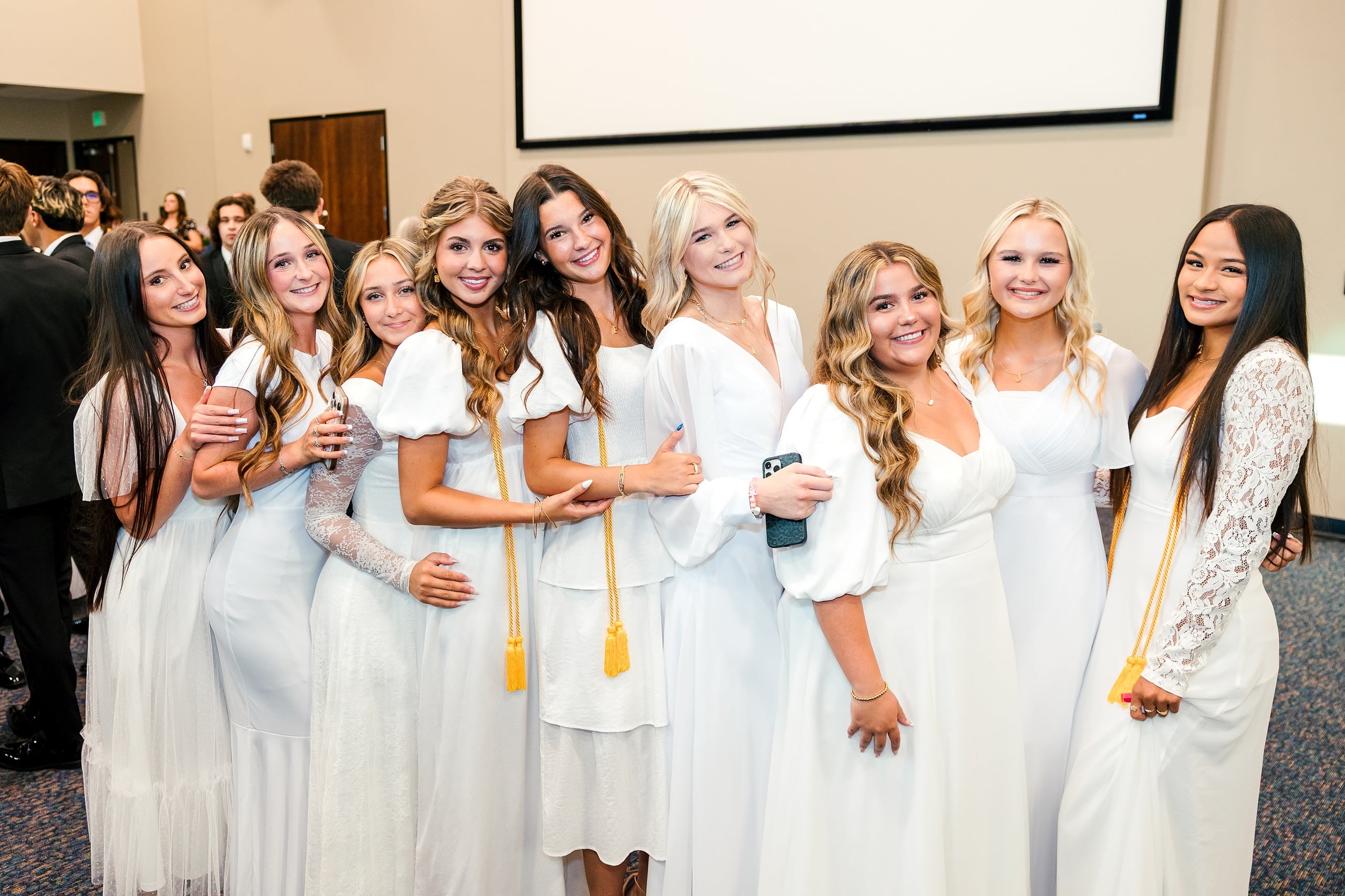 Christian High School Commencement 2025: Rise Up — 06 Female graduates in white dresses with gold cords gather together smiling