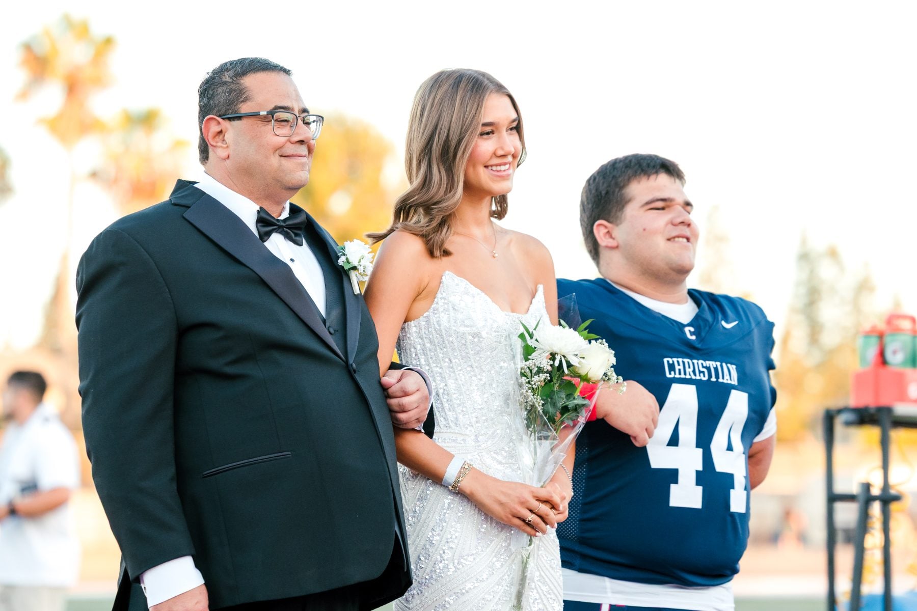 Homecoming court student in white dress poses with father and football player