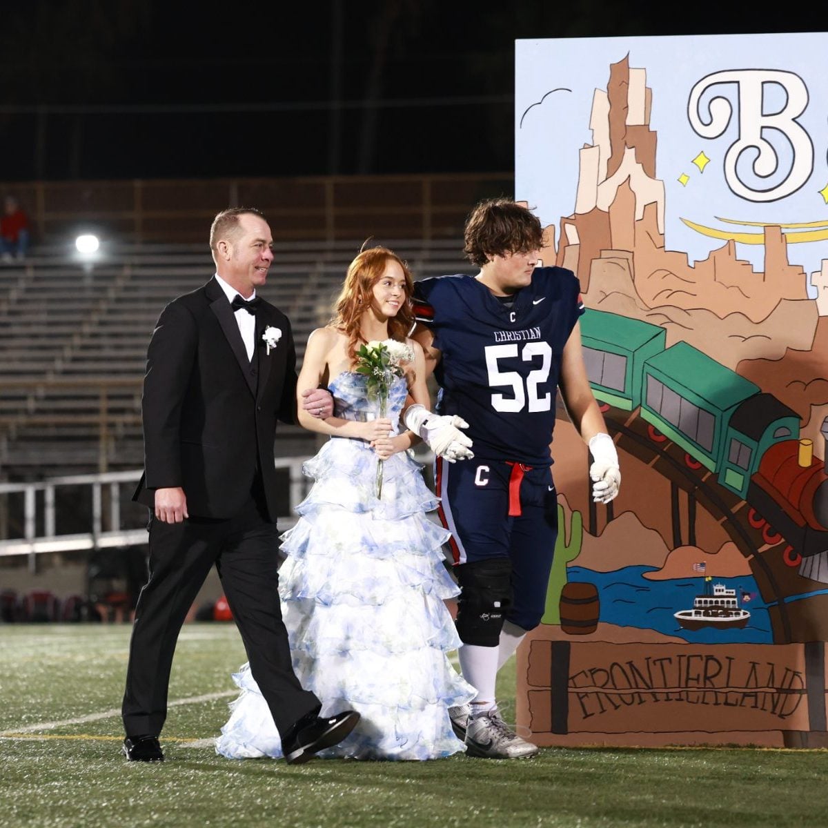 Father walks a student in white gown across the field, Patriots football player follows