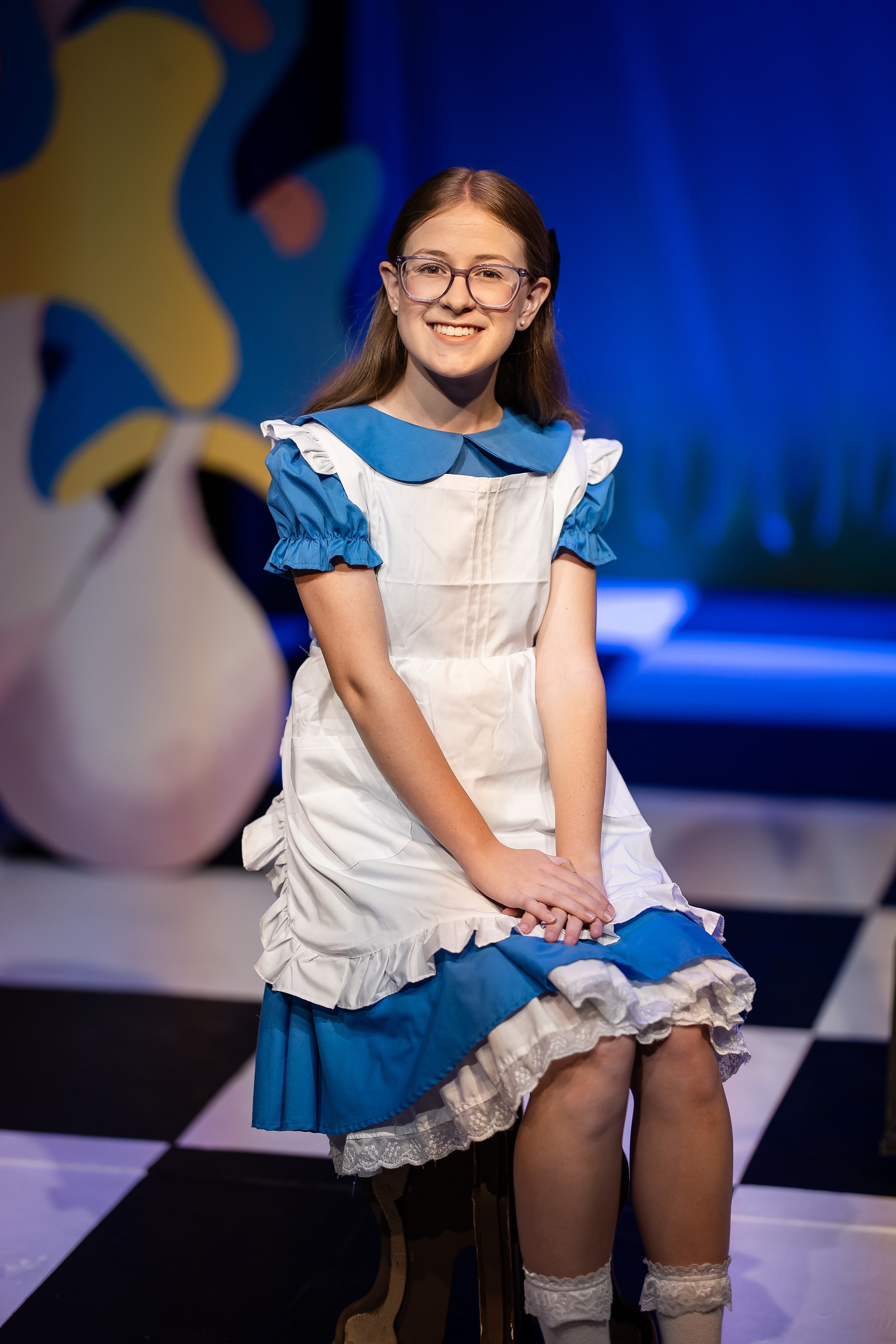 Curiouser and Curiouser! A Whimsical Journey with the Patriot Players — 02 Student portraying Alice with glasses sits on whimsical stage