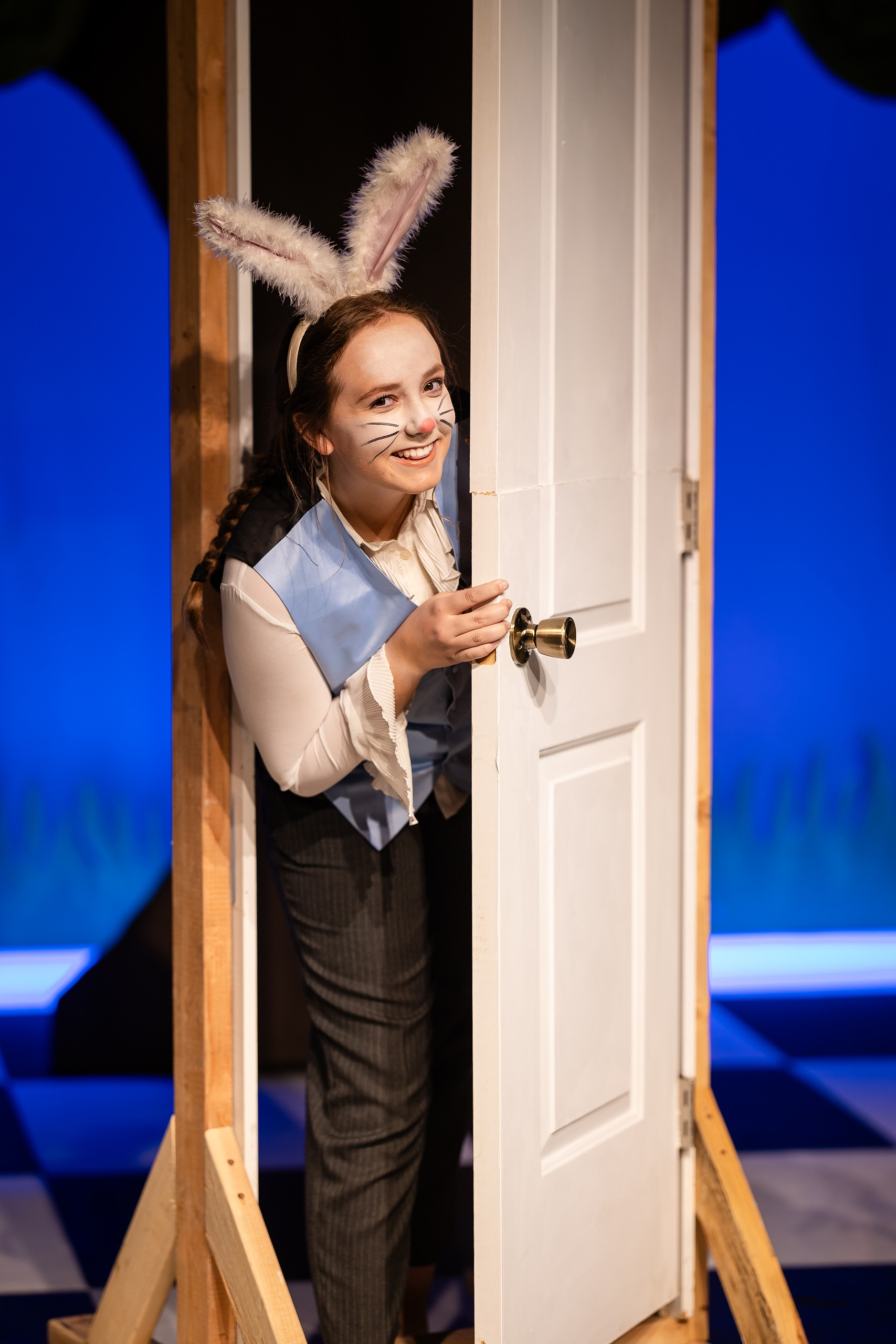 Curiouser and Curiouser! A Whimsical Journey with the Patriot Players — 05 White Rabbit character with bunny ears peers around white door on stage