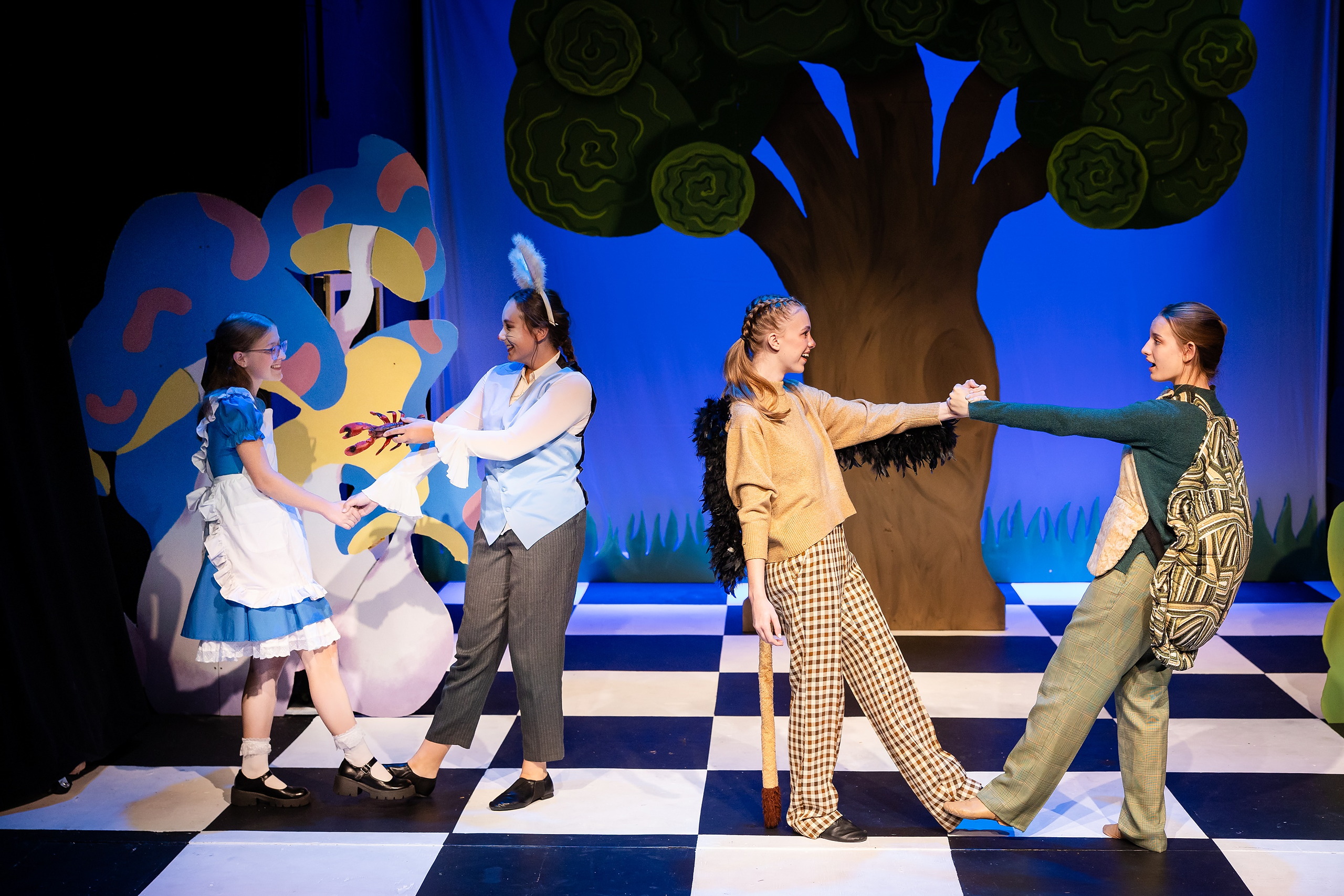 Curiouser and Curiouser! A Whimsical Journey with the Patriot Players — 17 Cast performs near large painted tree with whimsical Wonderland backdrop