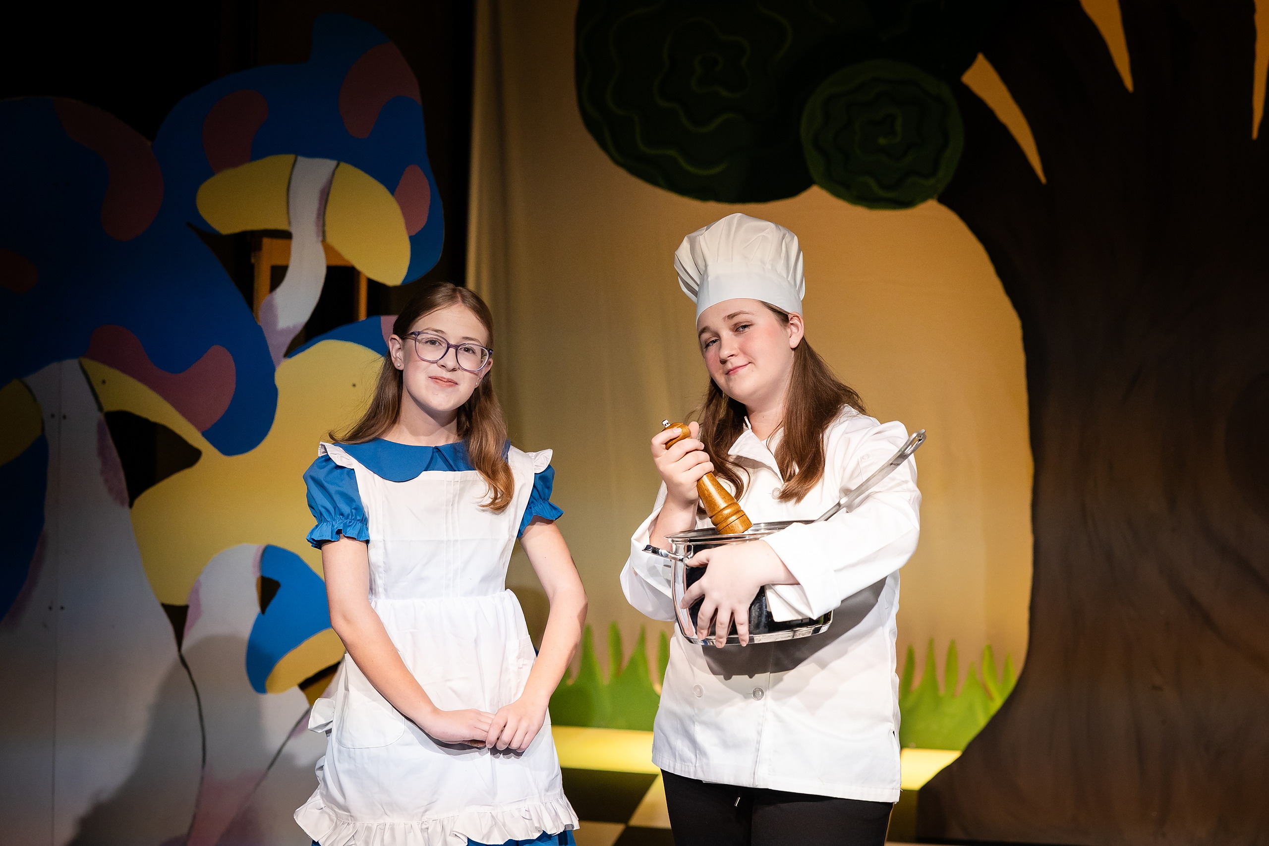 Curiouser and Curiouser! A Whimsical Journey with the Patriot Players — 18 Cook character in white stands with student in Alice costume on stage