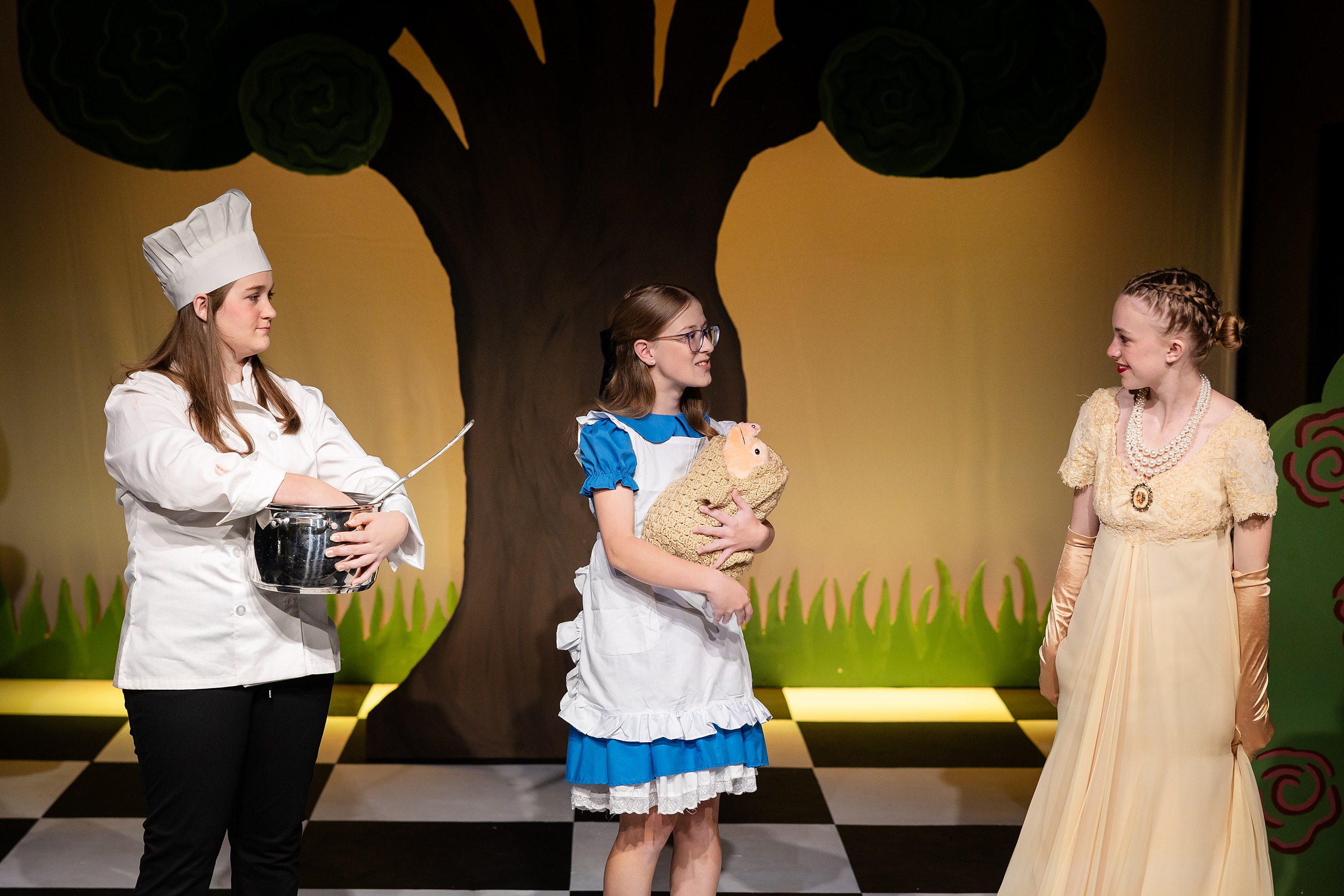Curiouser and Curiouser! A Whimsical Journey with the Patriot Players — 19 Cook, Alice, and a student in peach gown interact during scene