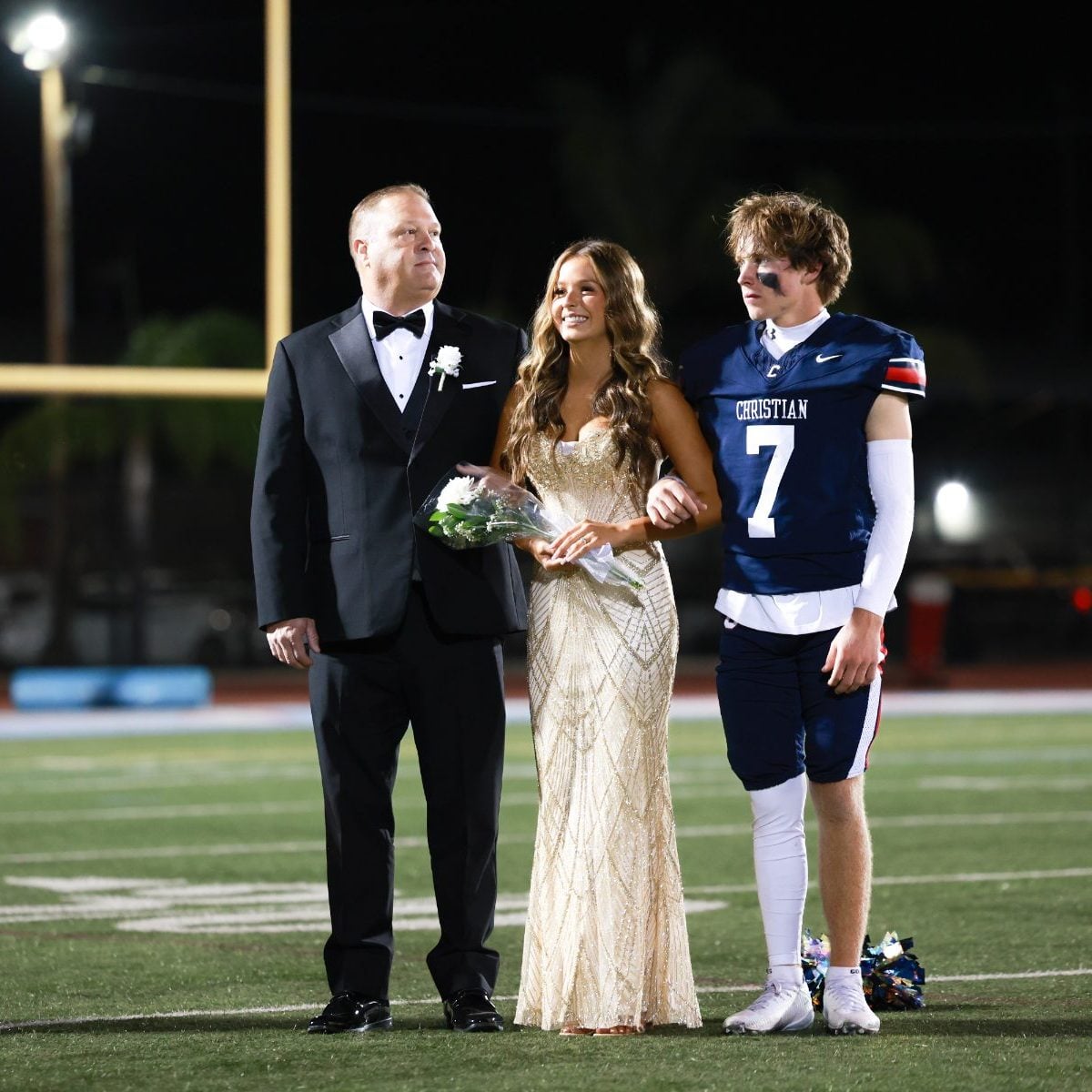 Homecoming court trio stands on the field, player wears jersey number 7