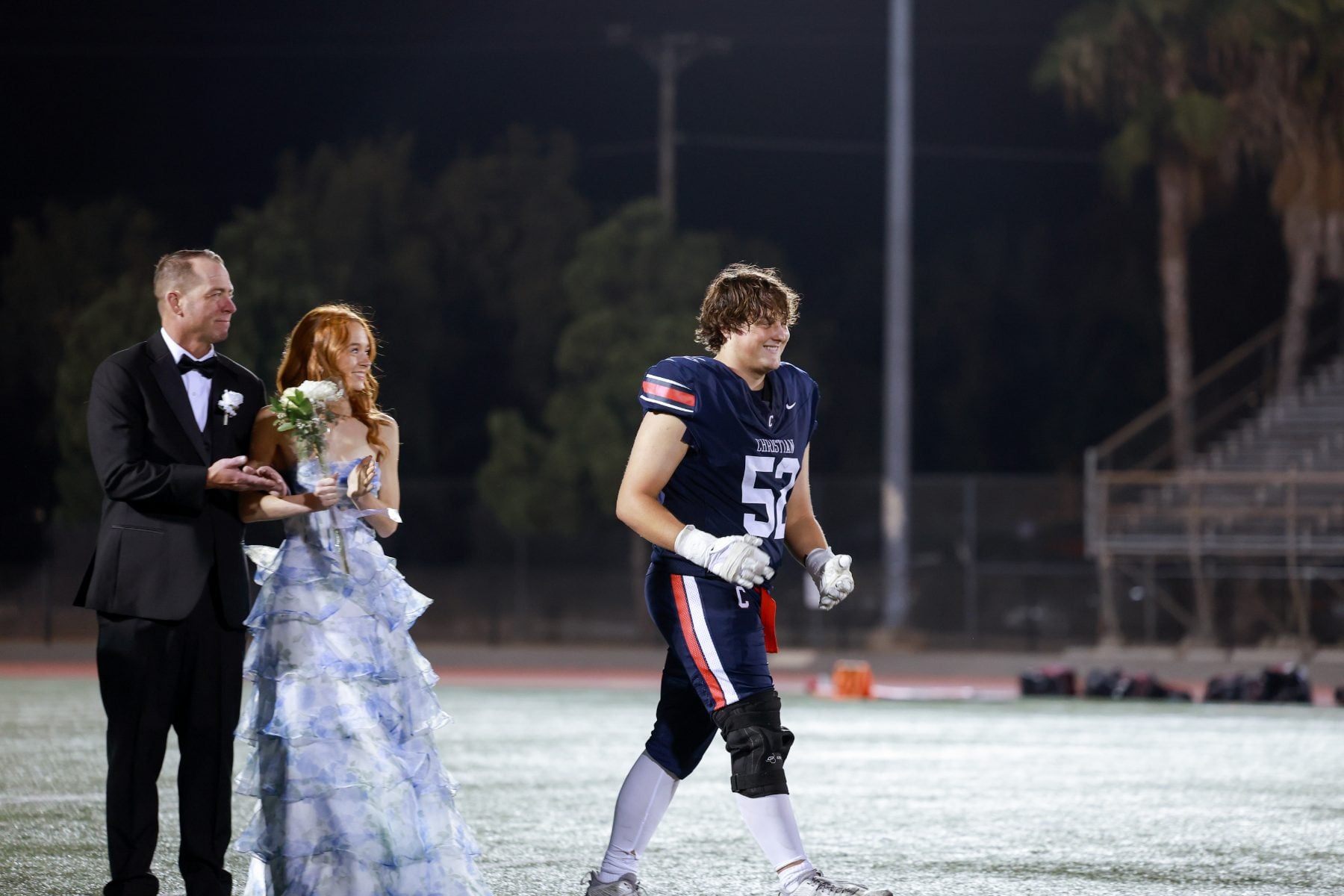 Football player number 52 walks past homecoming court couple on field