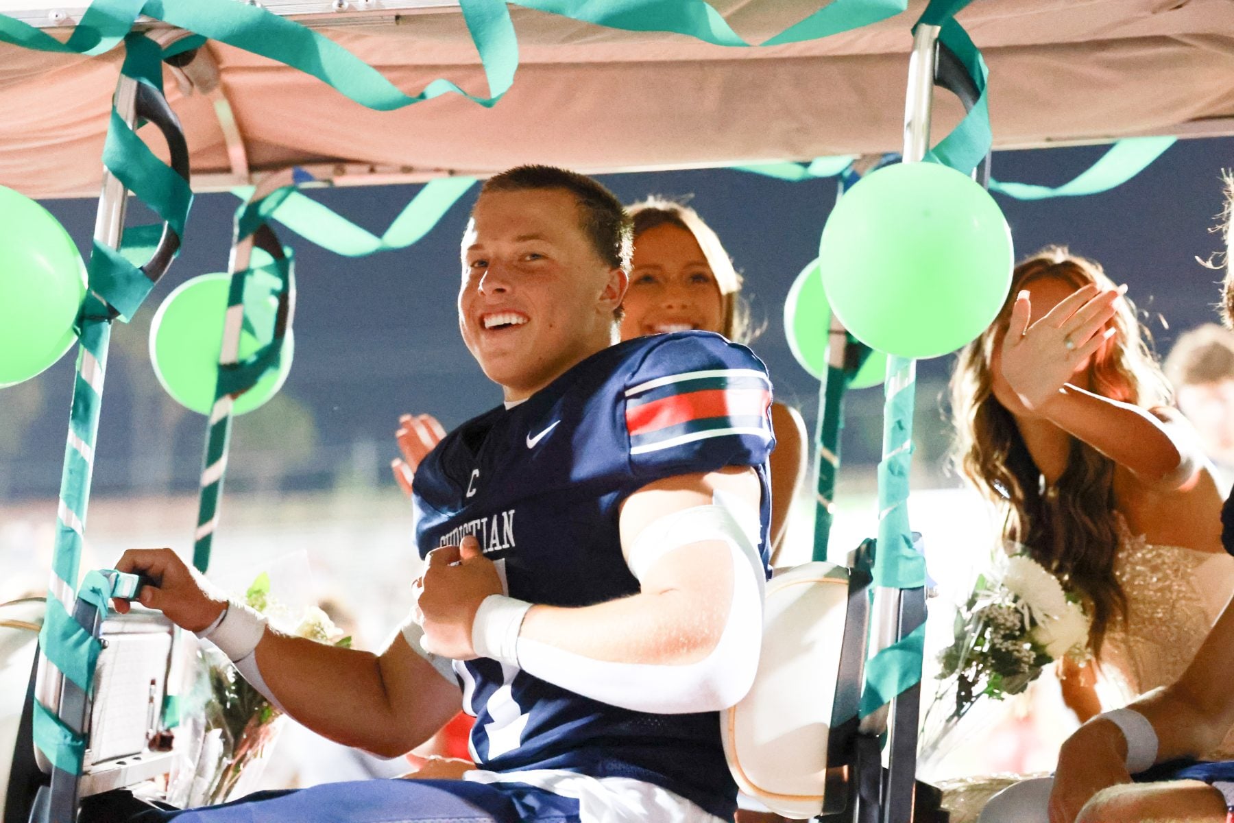 Football player smiles riding in homecoming parade cart with green balloons