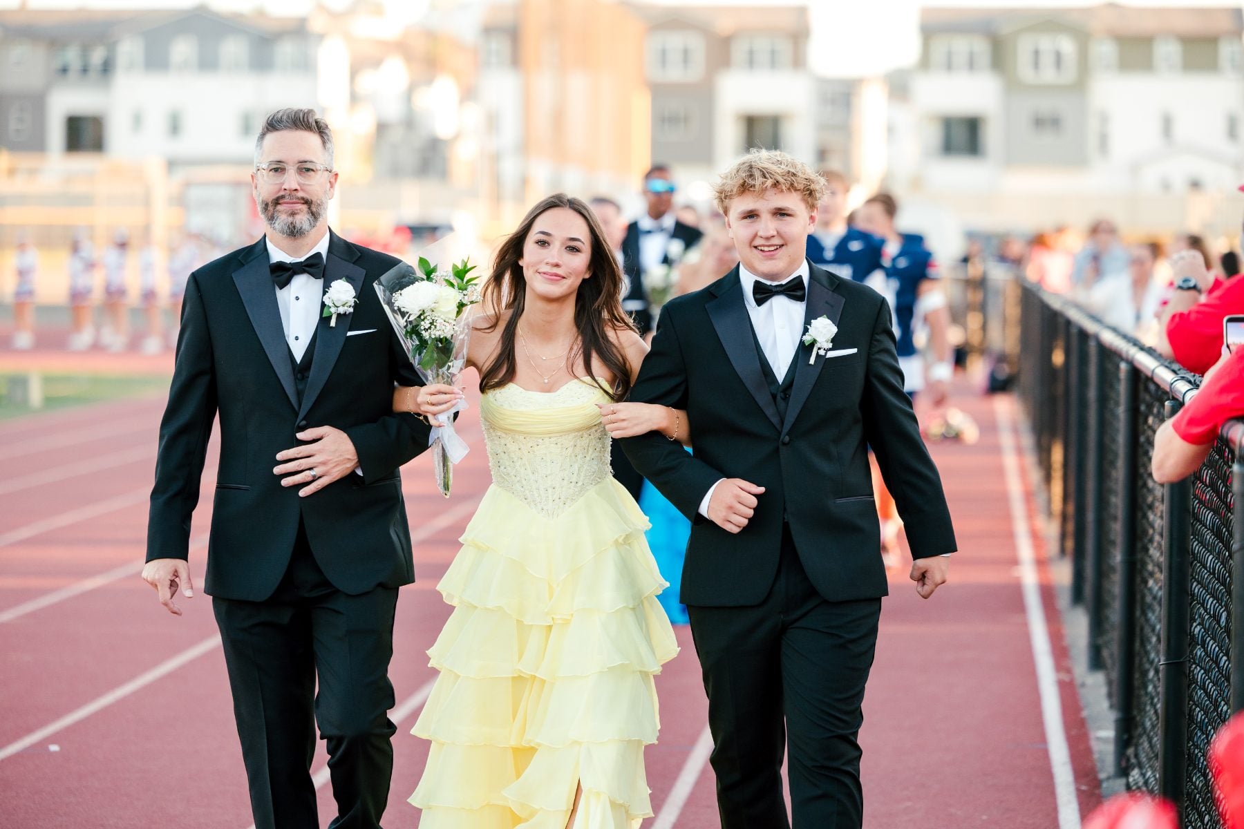 Homecoming queen candidate walks track escorted by two men in tuxedos