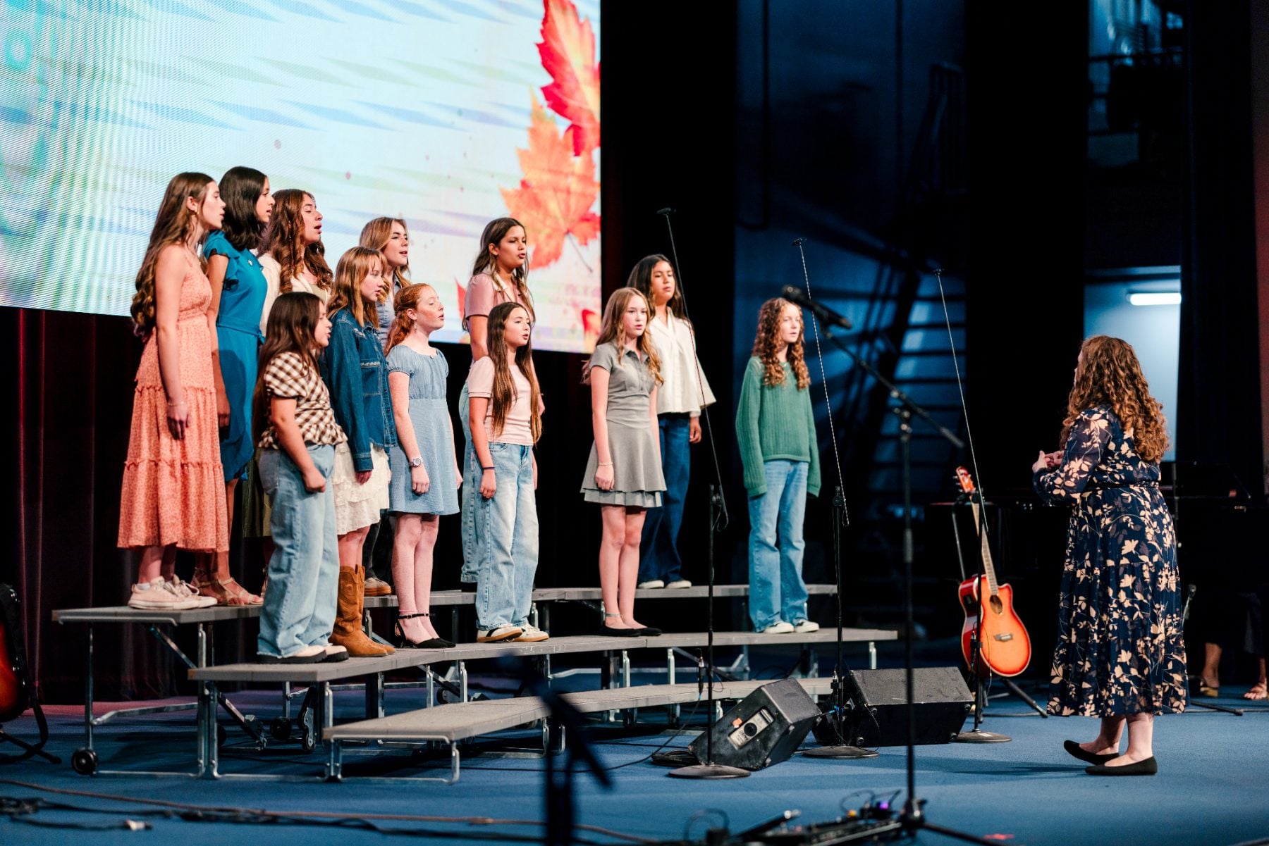 Young choir students sing while a teacher conducts on stage.